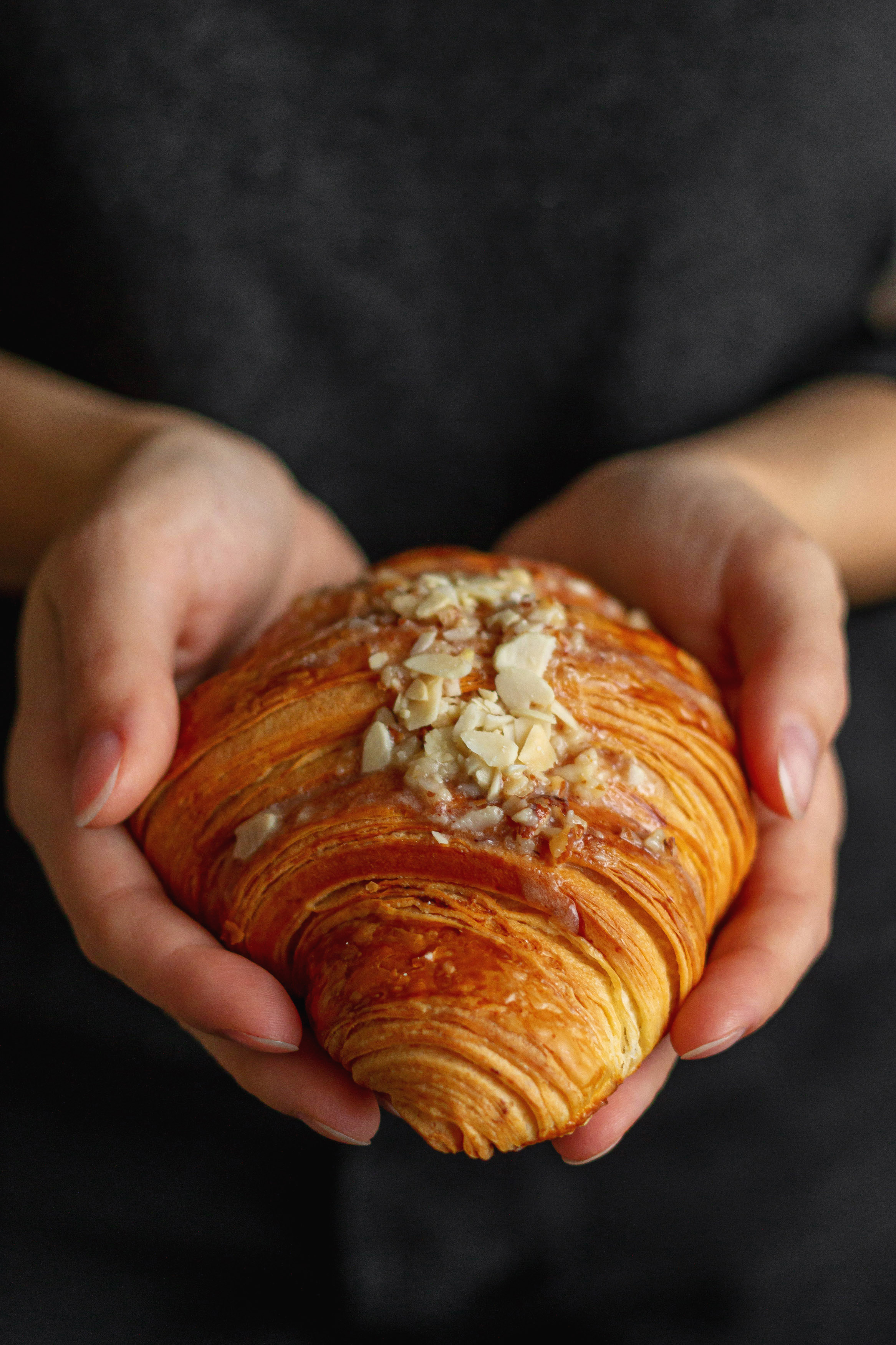 Женские руки держат выпечку в виде круассана на фоне черного фартука со светом от окна