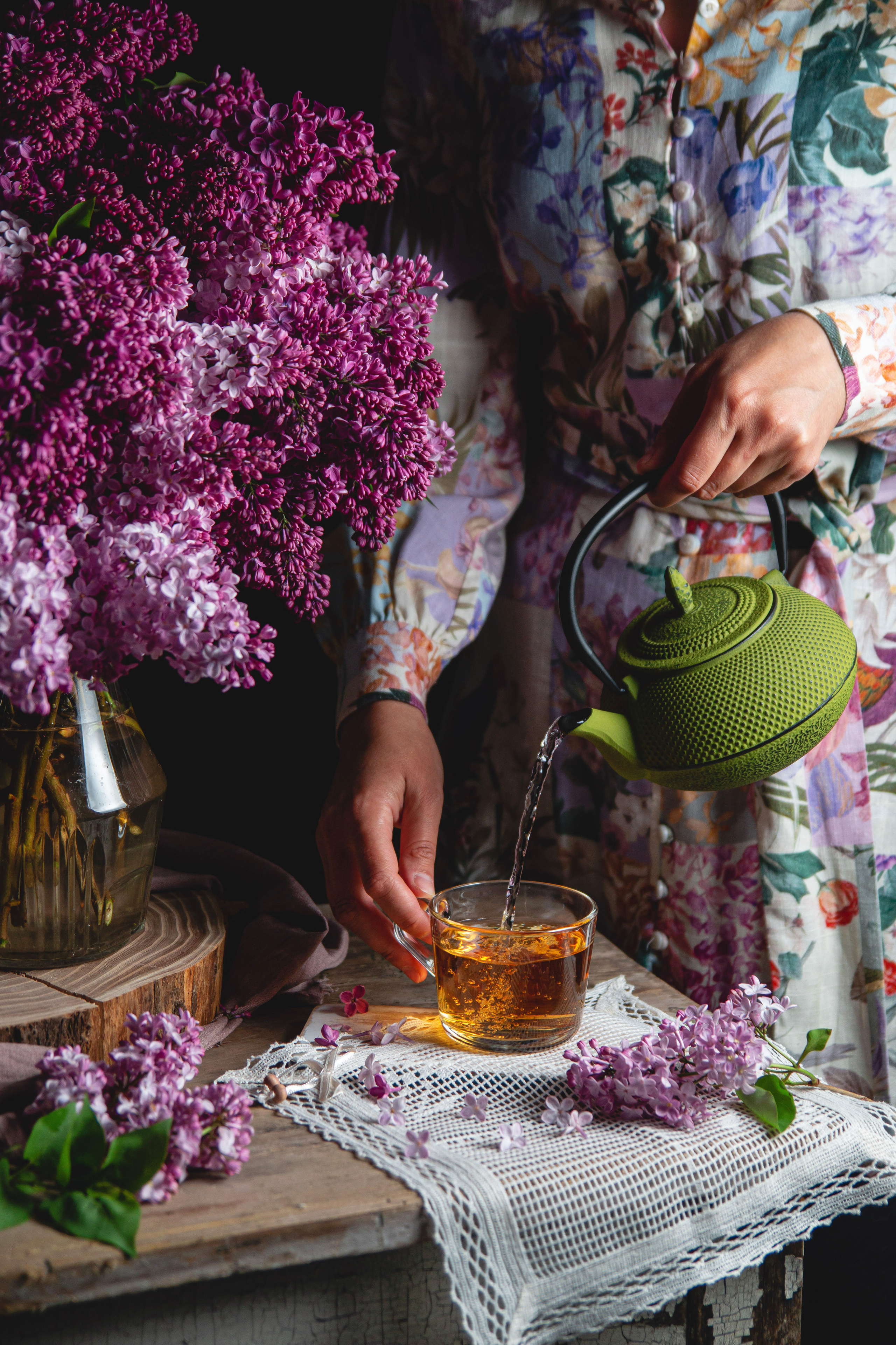 Проект сирень, девушка наливает чай в стакан из зеленого чугунного чайника на фоне сирени и винтажного деревянного стола со светом от окна