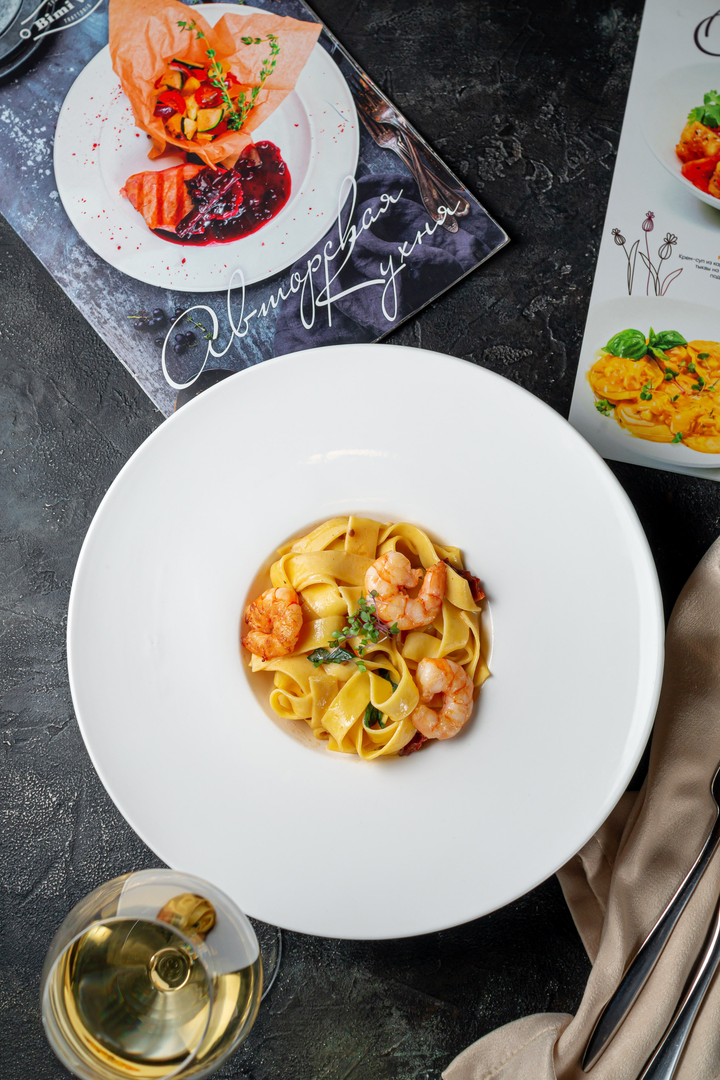 Паста с креветками в тарелке с широкими краями стоит на столе из темного камня, рядом меню из ресторана Bimi Vita и салфетка со столовыми приборами