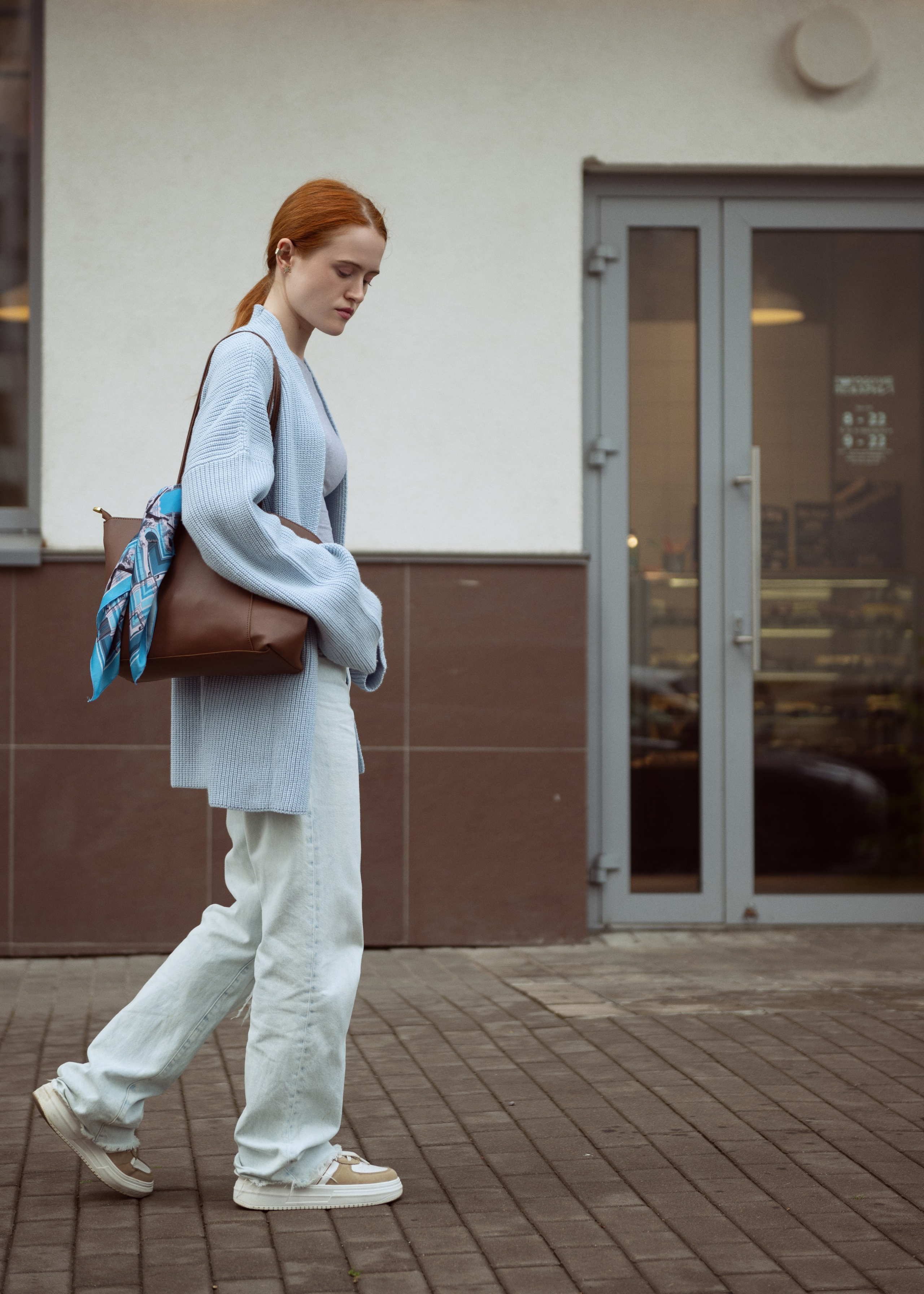 Красивая рыжеволосая девушка в голубом кардигане и джинсах идет по улице, на плече у нее коричневая сумка, украшенная голубым шелковым платком.