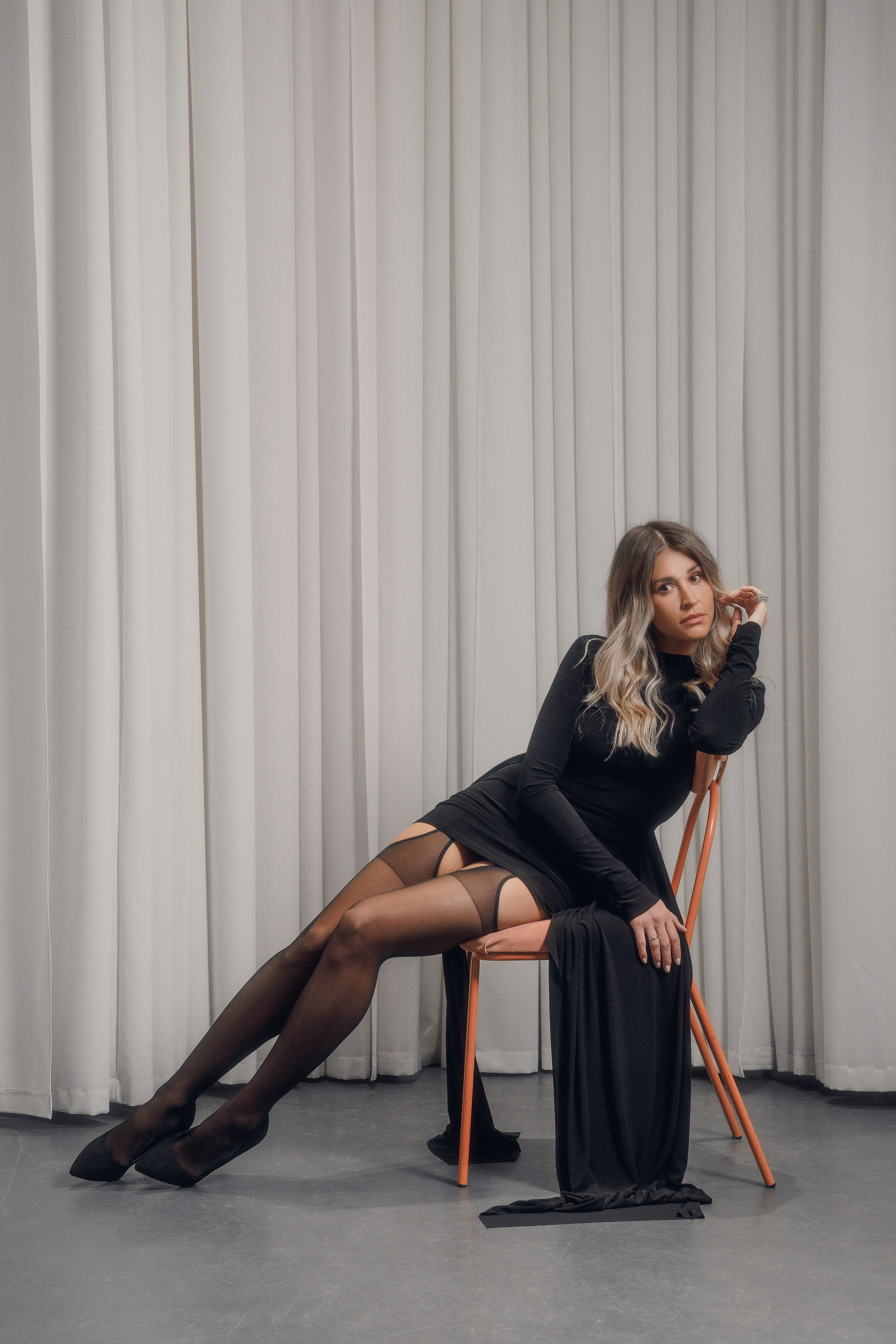 Модный портрет: девушка в черном платье на стуле перед занавесками.