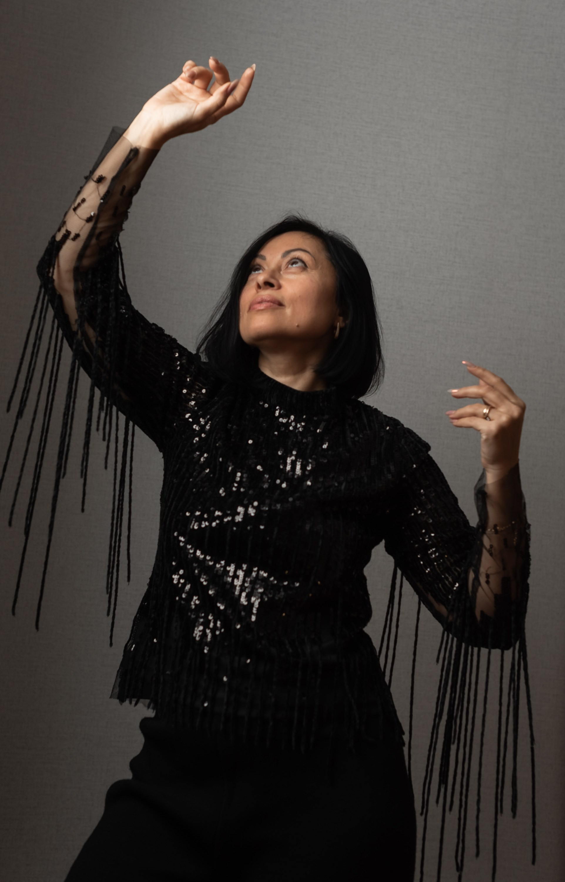 Девушка в блузке с пайетками танцует танец, подняла руки и смотрит вверх.