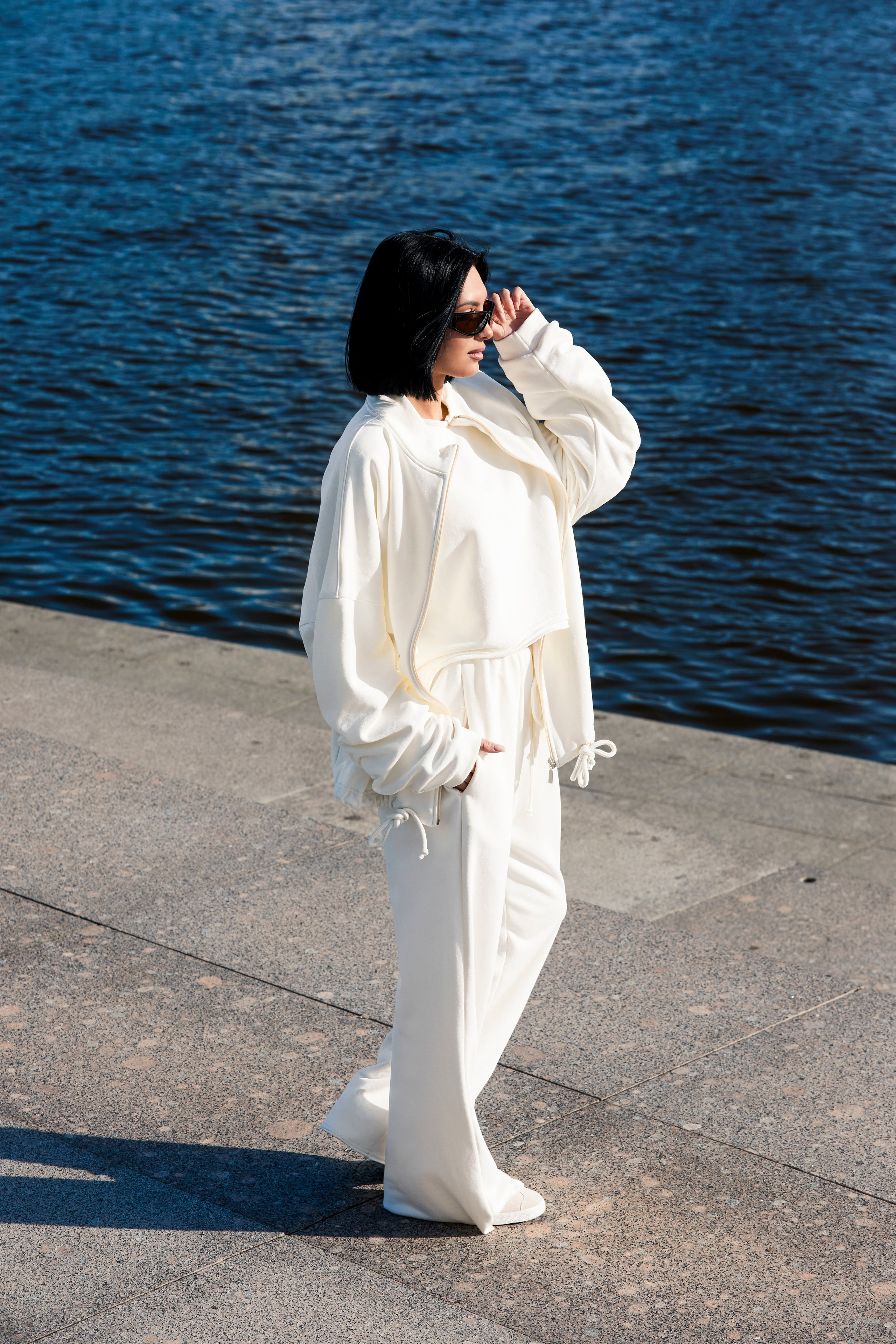 Девушка брюнетка в белом костюме и солнцезащитных очках стоит на фоне воды