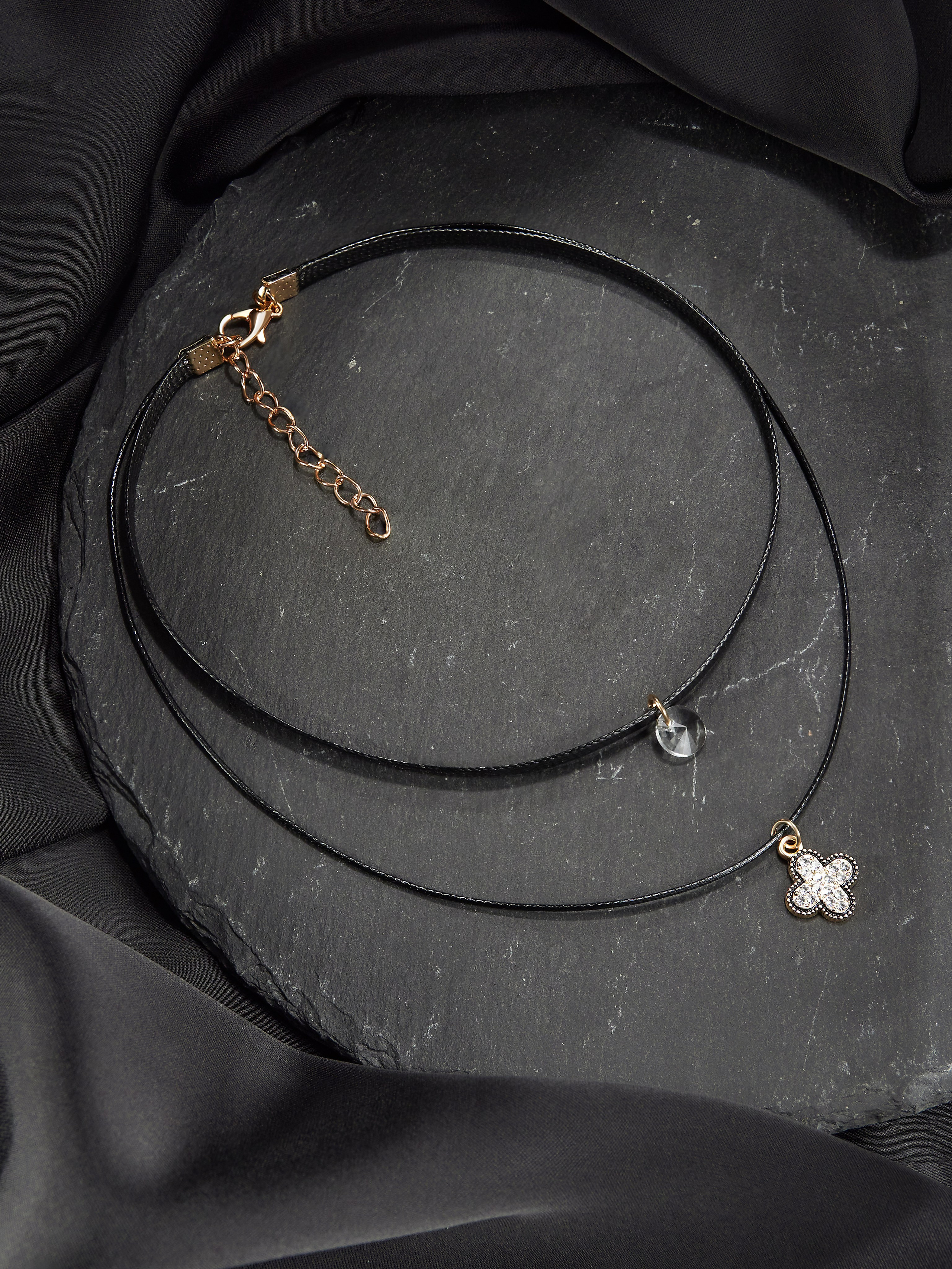 Черное двухрядное колье-чокер с золотой застежкой-цепочкой и регулируемым удлинителем лежит на черной фактурной поверхности. На одной нити изображен небольшой кулон с белым цветком, а на другой — драгоценный камень в форме капли. Фон украшен черной шелковистой тканью.