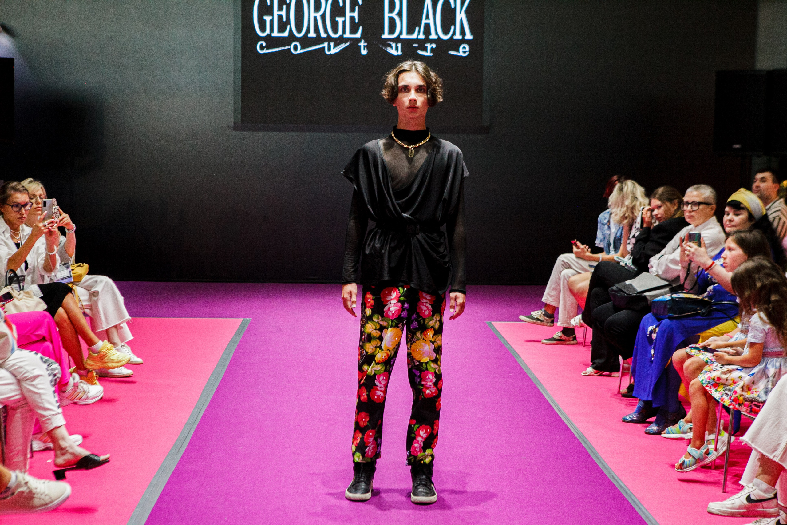  "Модели в ярких нарядах проходят по подиуму во время показа мод, освещенные яркими софитами и окруженные восторженными зрителями."