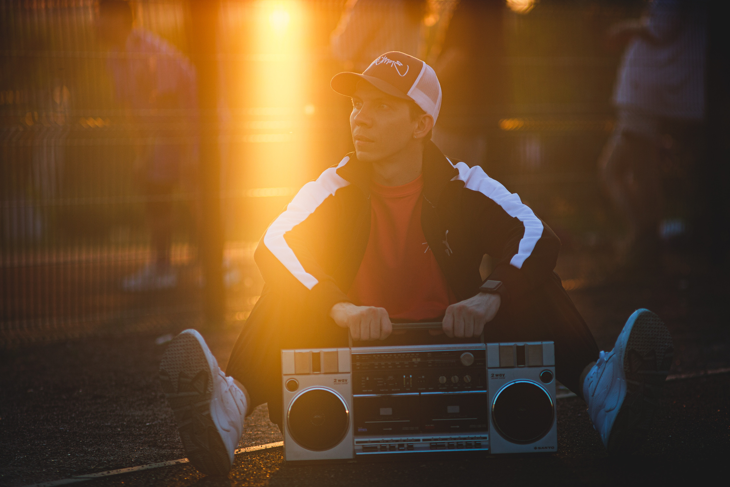 Парень в спортивной костюме в бейсболке с магнитофоном сидит на фоне спортивной площадки в лучах закатного солнца