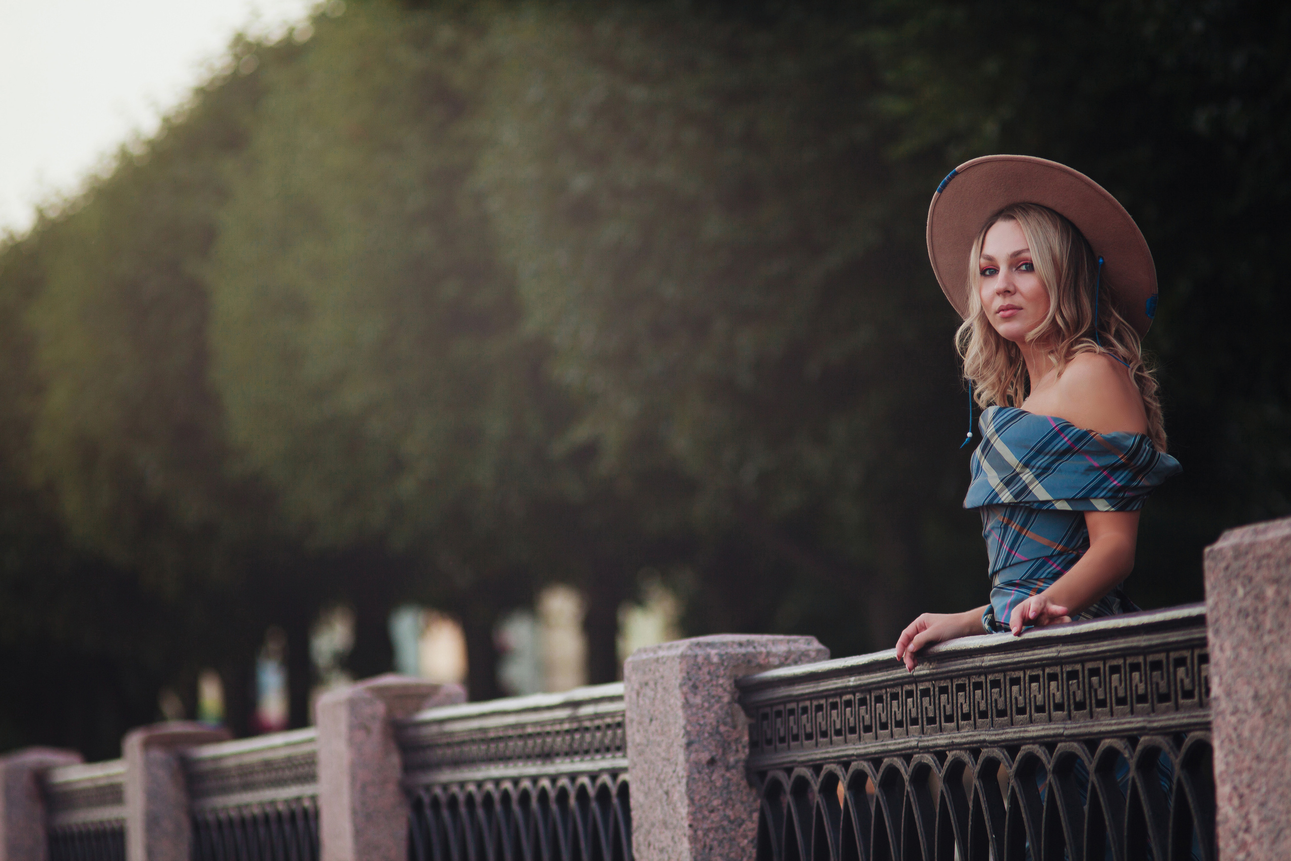 Девушка в шляпе стоит у парапета и зеленых деревьев