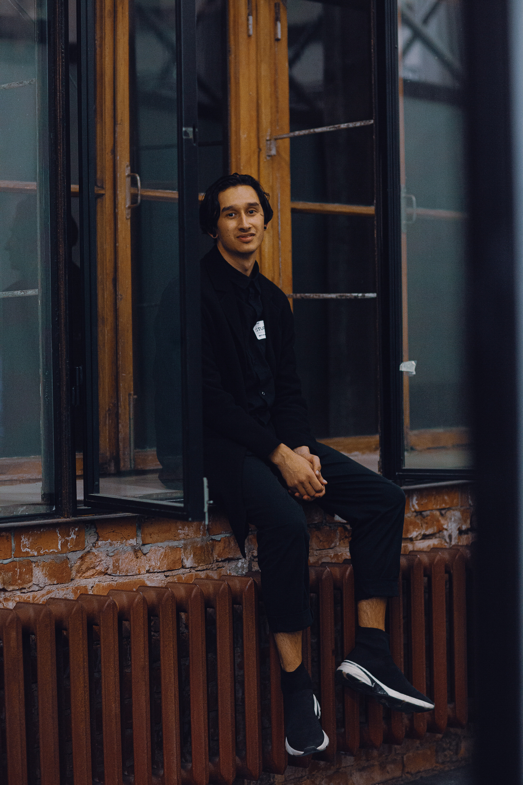 Молодой человек в черной одежде и кроссовках в помещении на фоне окон