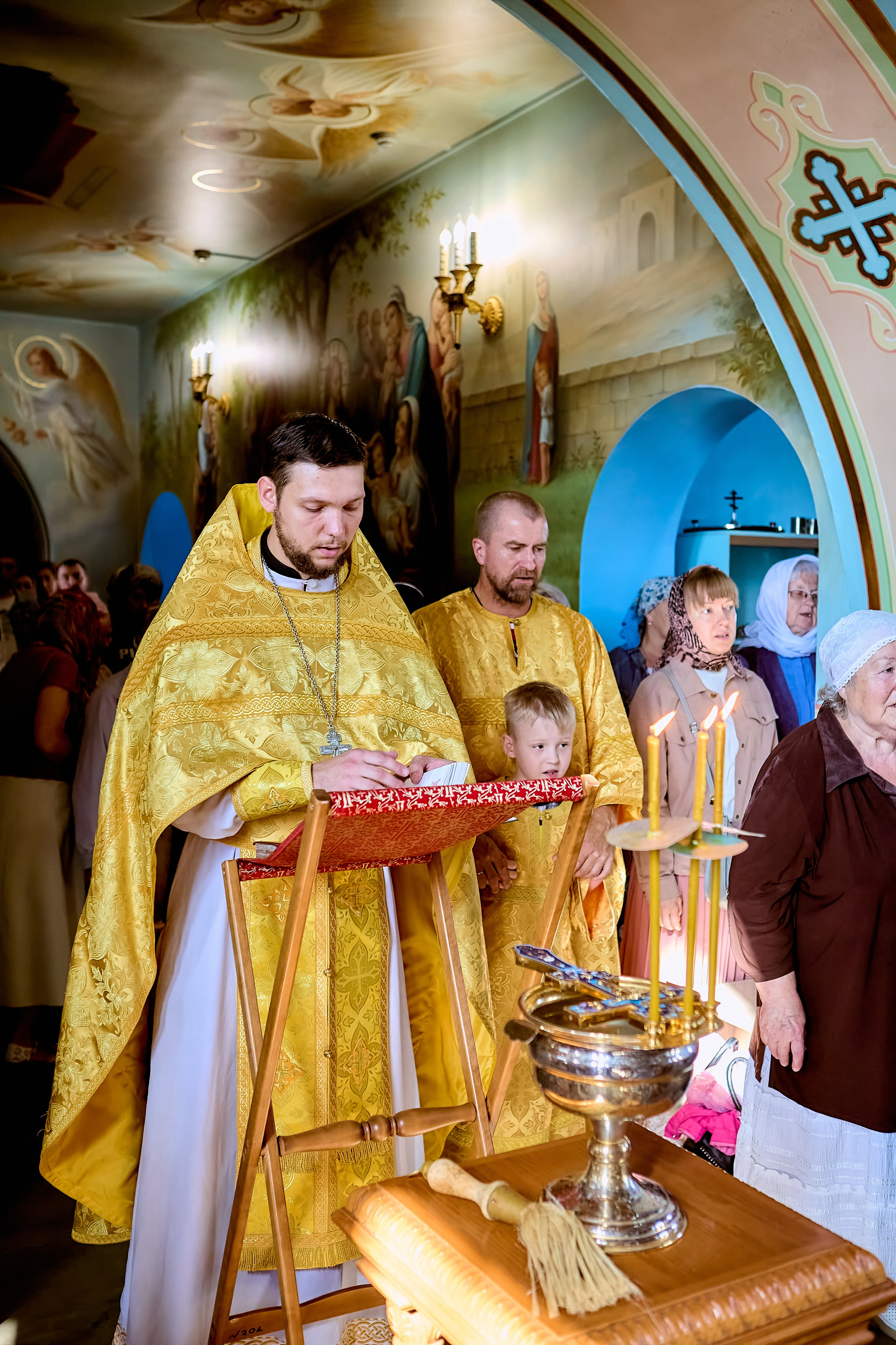 Священник стоит у аналое и читает молитвы для освещения воды на столике стоит чаша с водой и горят свечи