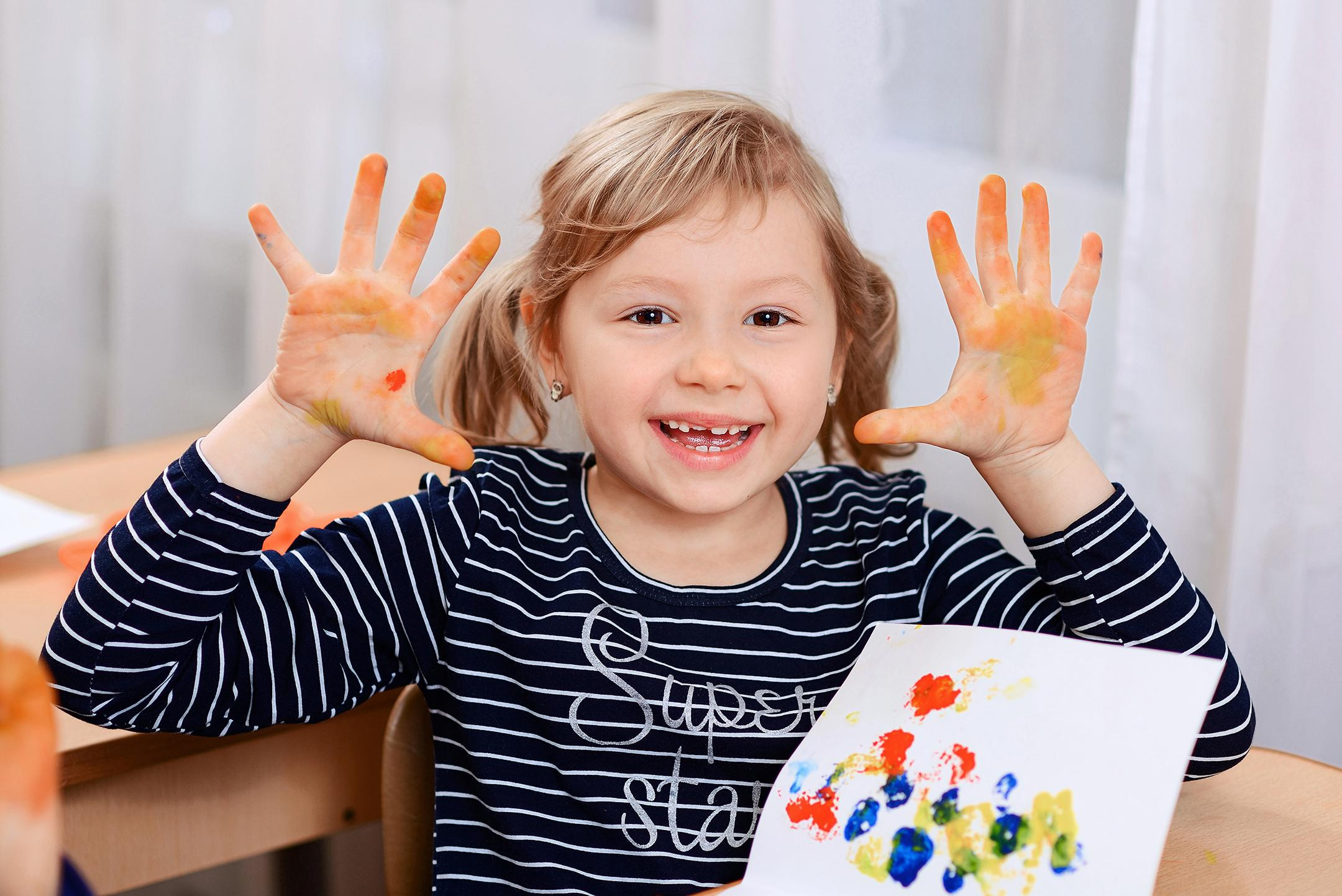 Девочка после рисования в детском саду показывает грязные от краски ладошки
