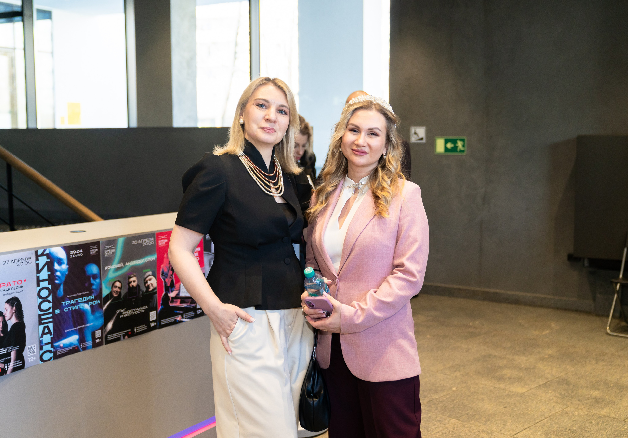 Участницы празднования Дня местного самоуправления в Санкт-Петербурге. Две женщины в деловых образах позируют у стенда с афишами. Фото Евгения Мальцева.