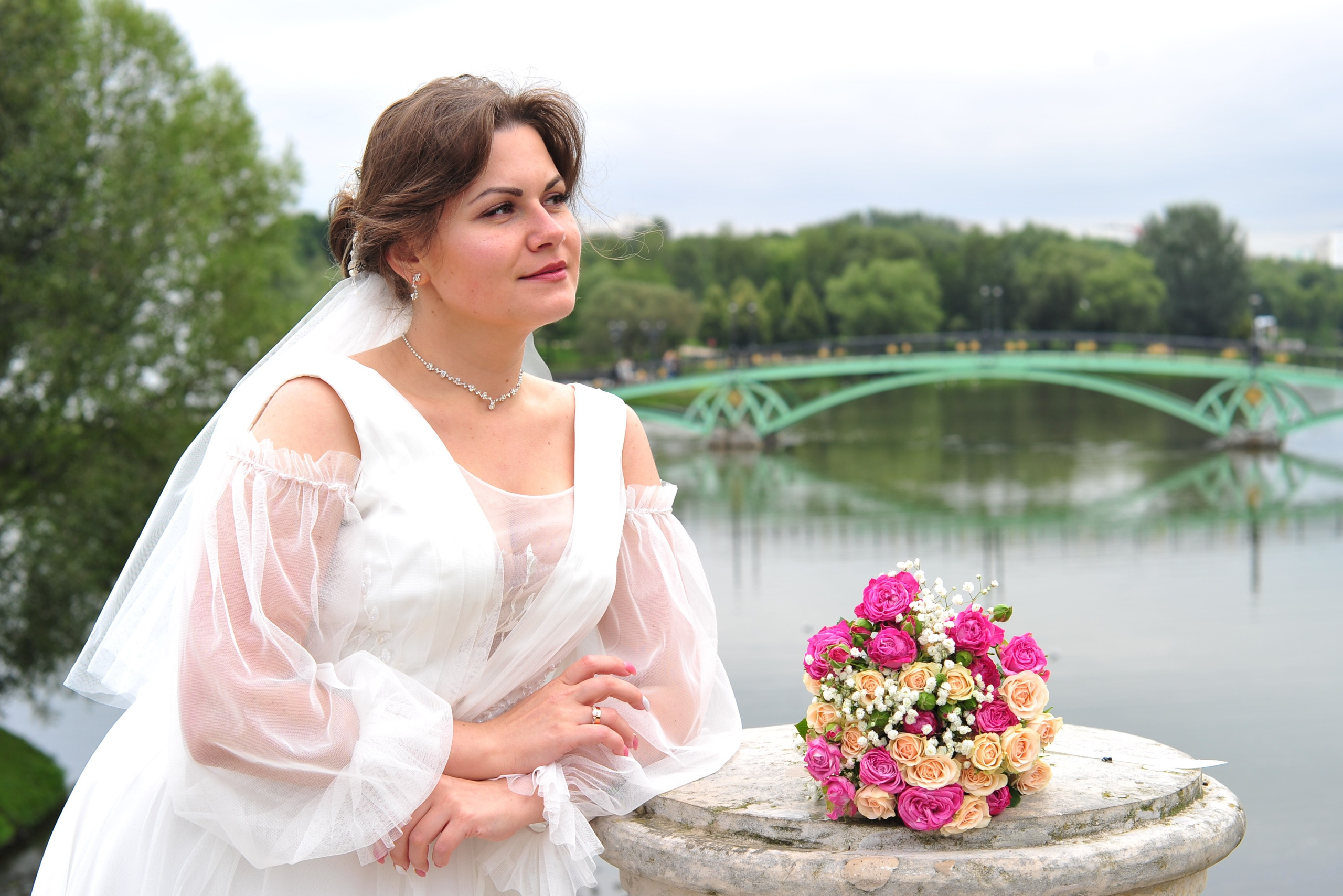 Свадьба в Царицыно пасмурным летним днем, невеста стоит на каменном мосту, букет невесты лежит на каменной колонне моста на фоне пруда и железного моста вдалеке 