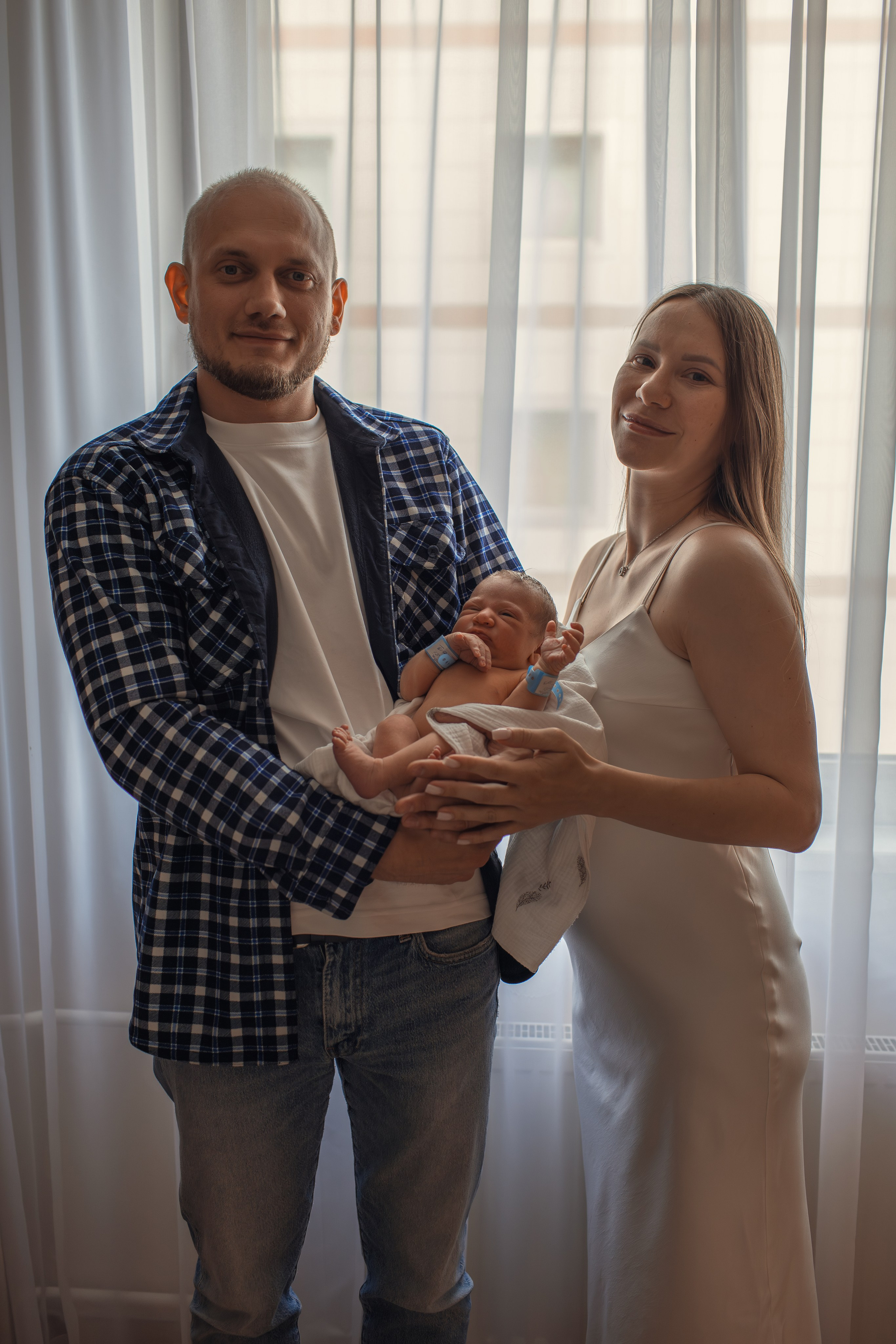 Родители вместе держат новорождённого малыша, улыбаясь в камеру.