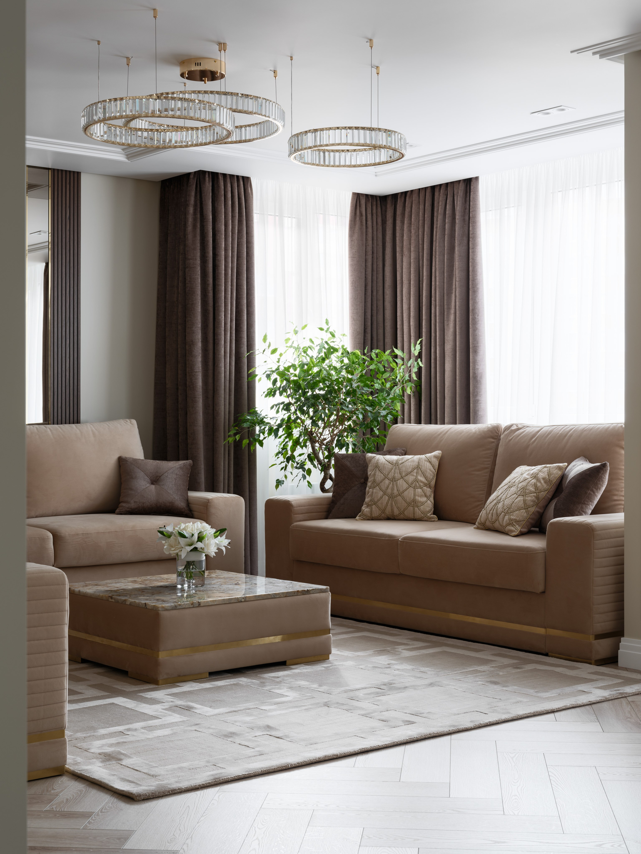 Стильная гостиная с мягкими бежевыми диванами, пышными шторами шоколадного оттенка, большим зелёным растением и журнальным столиком из камня с вазой белых цветов в центре интерьера.