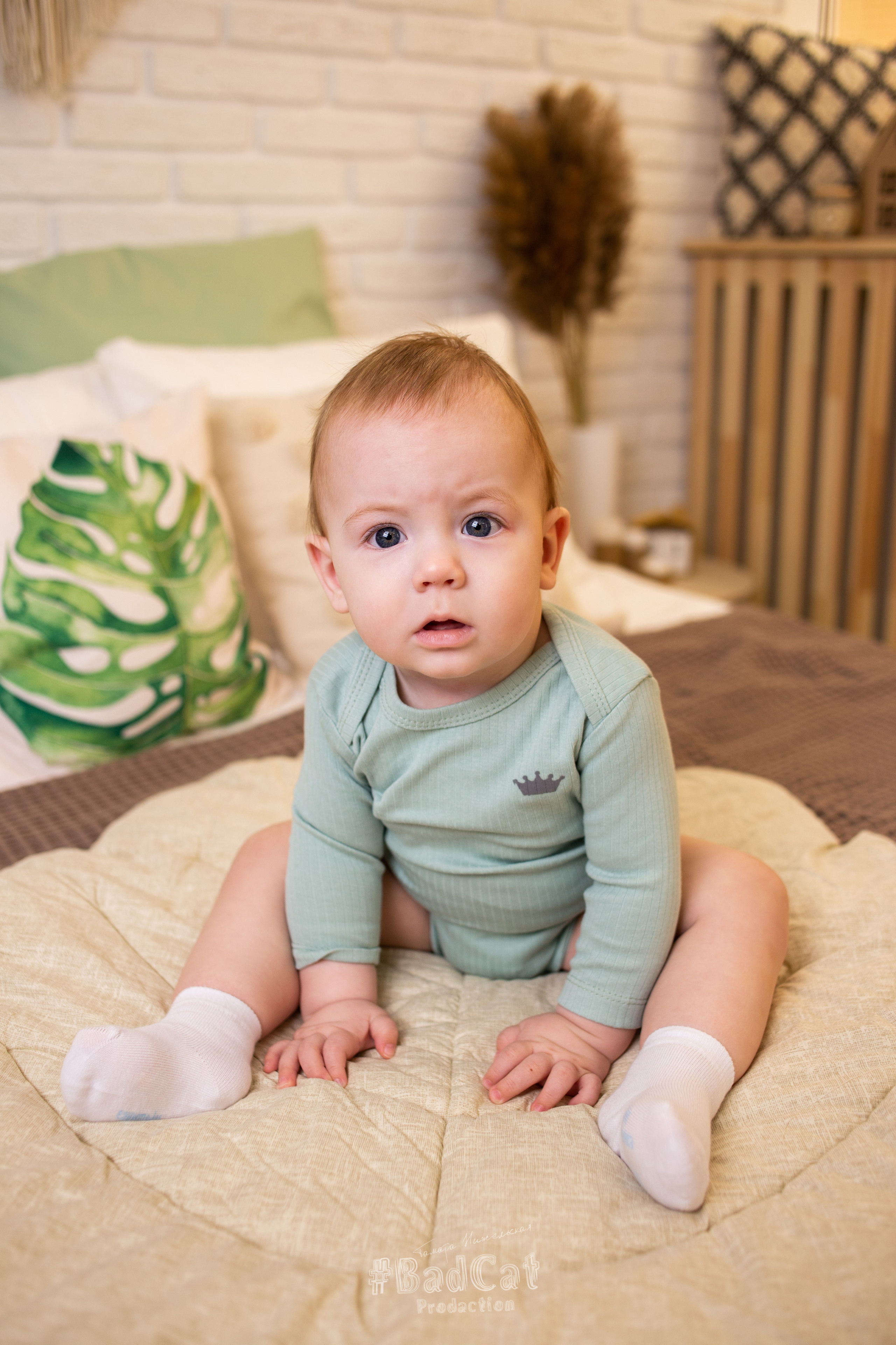 ребенок  в  зеленом  боди сидит на  бежевой  кровати