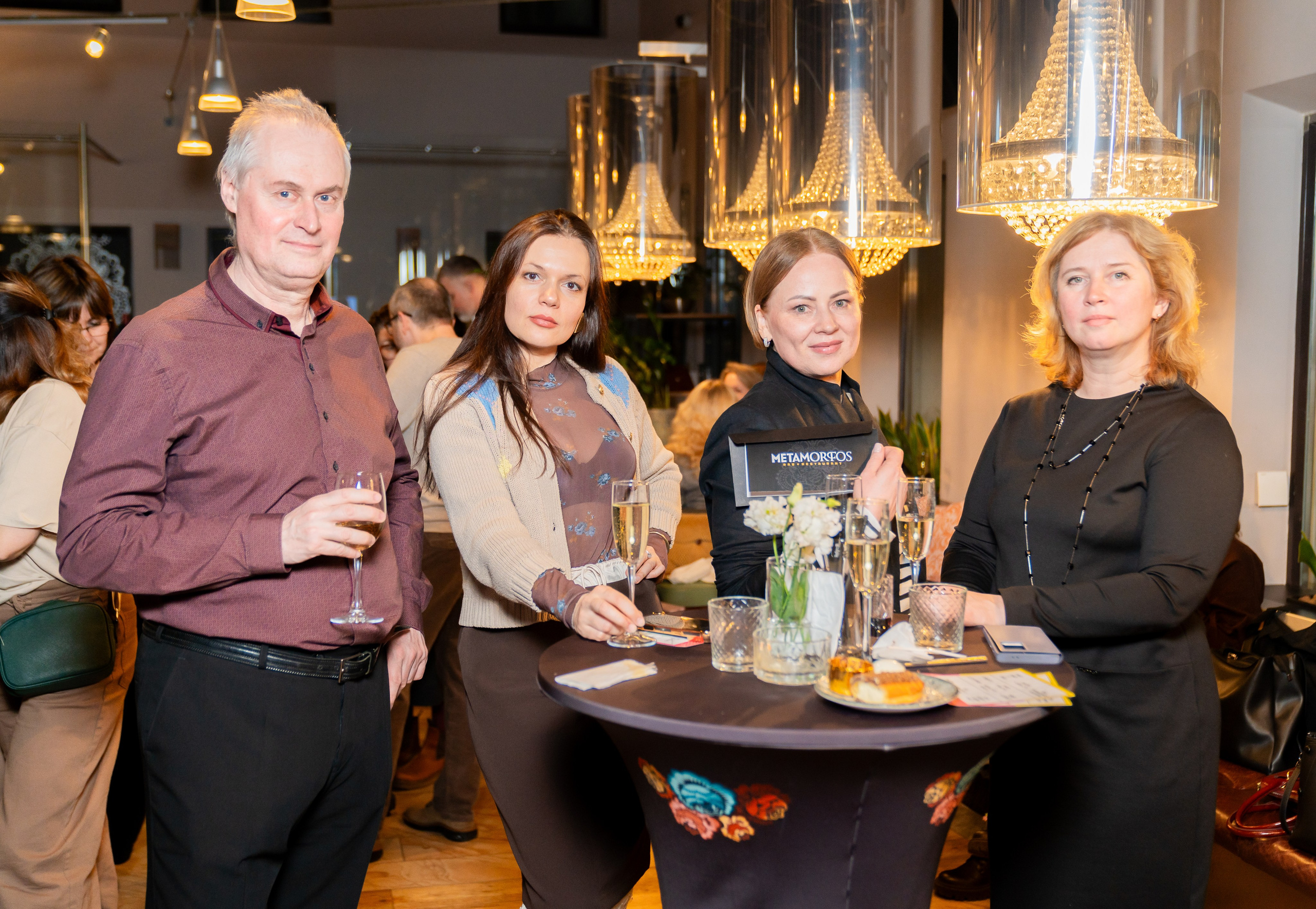 Мужчина в бордовой рубашке и три женщины позируют с бокалами шампанского на корпоративном мероприятии в Radisson Sonya. Фотография Евгения Мальцева (evrednix.ru).