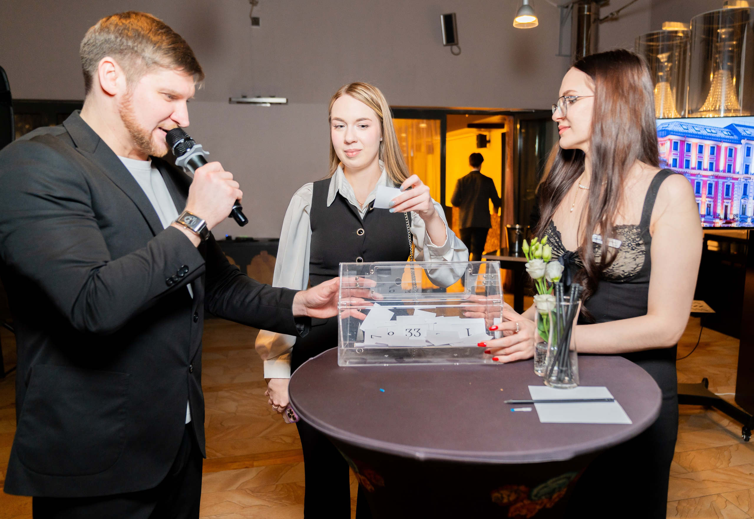 Ведущий с микрофоном проводит розыгрыш призов у прозрачного бокса на корпоративном мероприятии в Radisson Sonya. Две девушки-организатора за столом. Фото Евгения Мальцева (evrednix.ru).