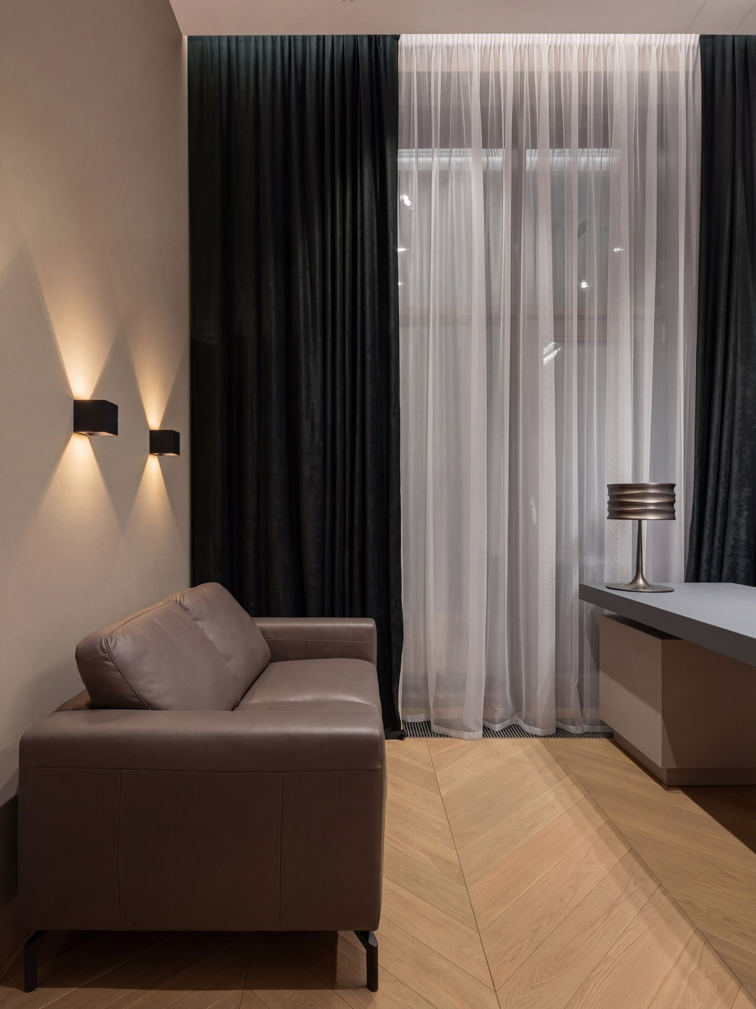 Интерьер кабинета от Валерии Котович: кожаный диван таупового цвета, графитовый рабочий стол, архитектурное освещение бра на стене и паркет «французская елка» в современном доме.