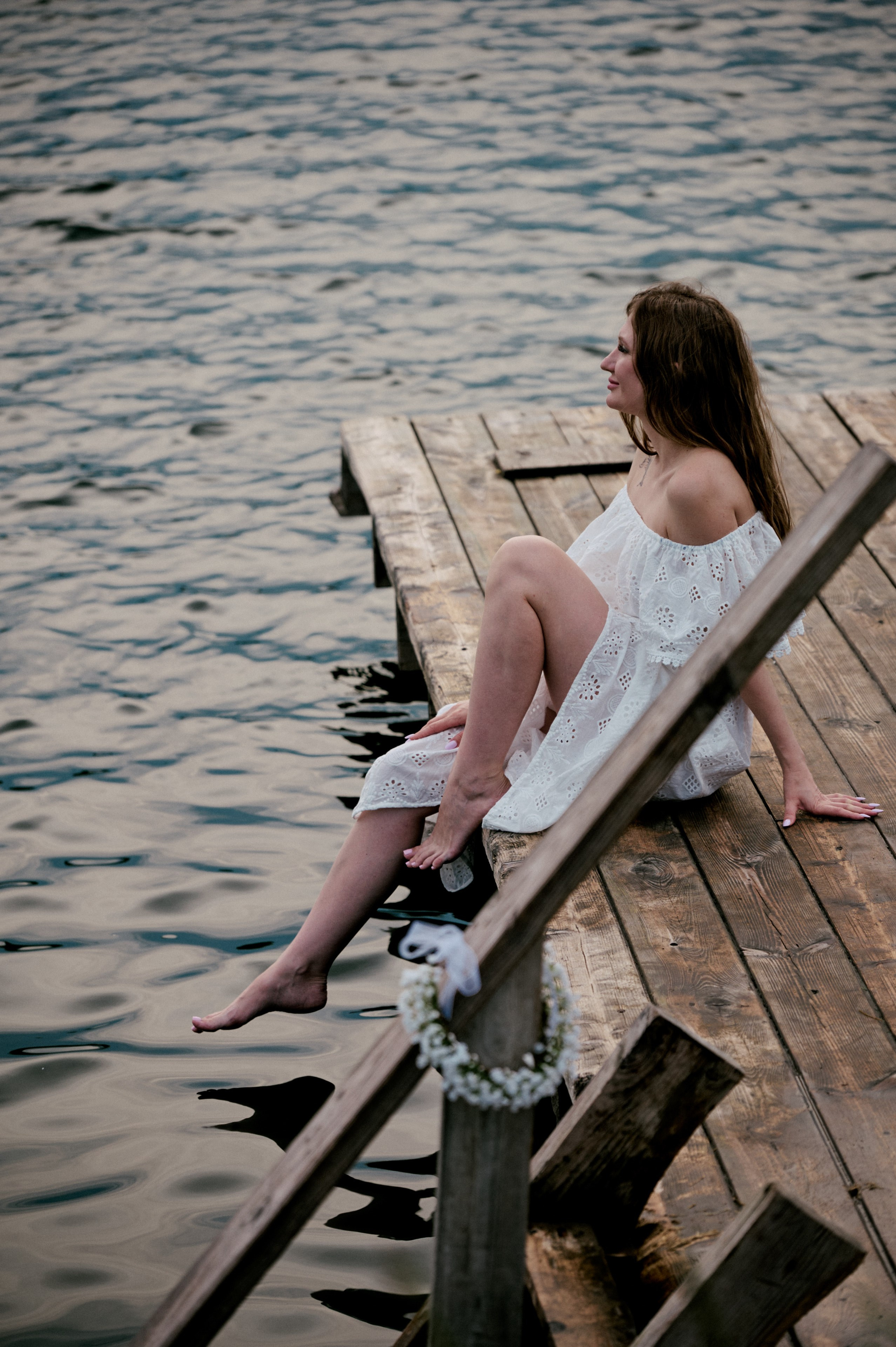 Евгения сидит на пирсе, свесив ноги над водой, в белом кружевном платье.
