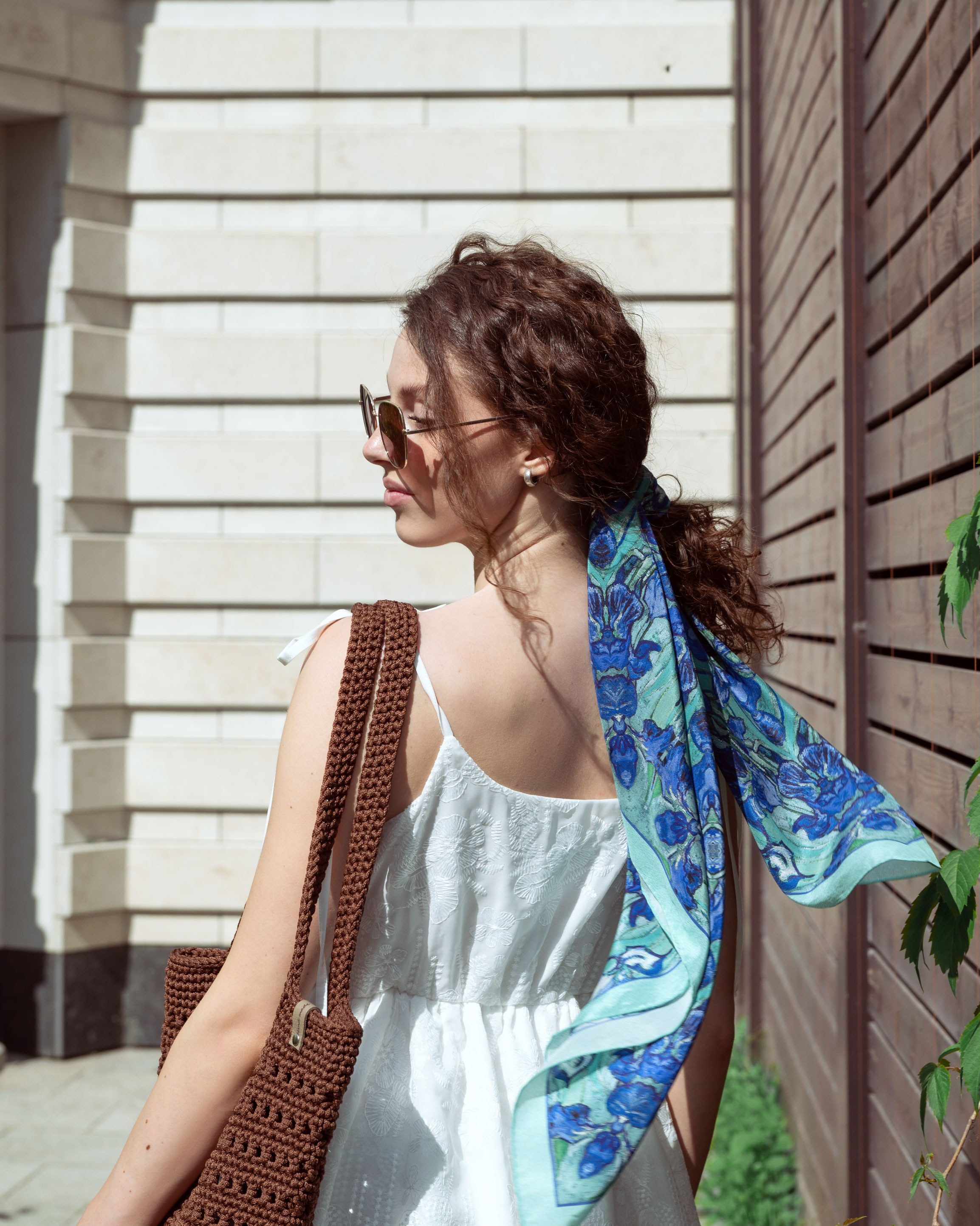 Красивая девушка в дизайнерском шелковом платке на волосах и в белом летнем платье идет вдоль стены