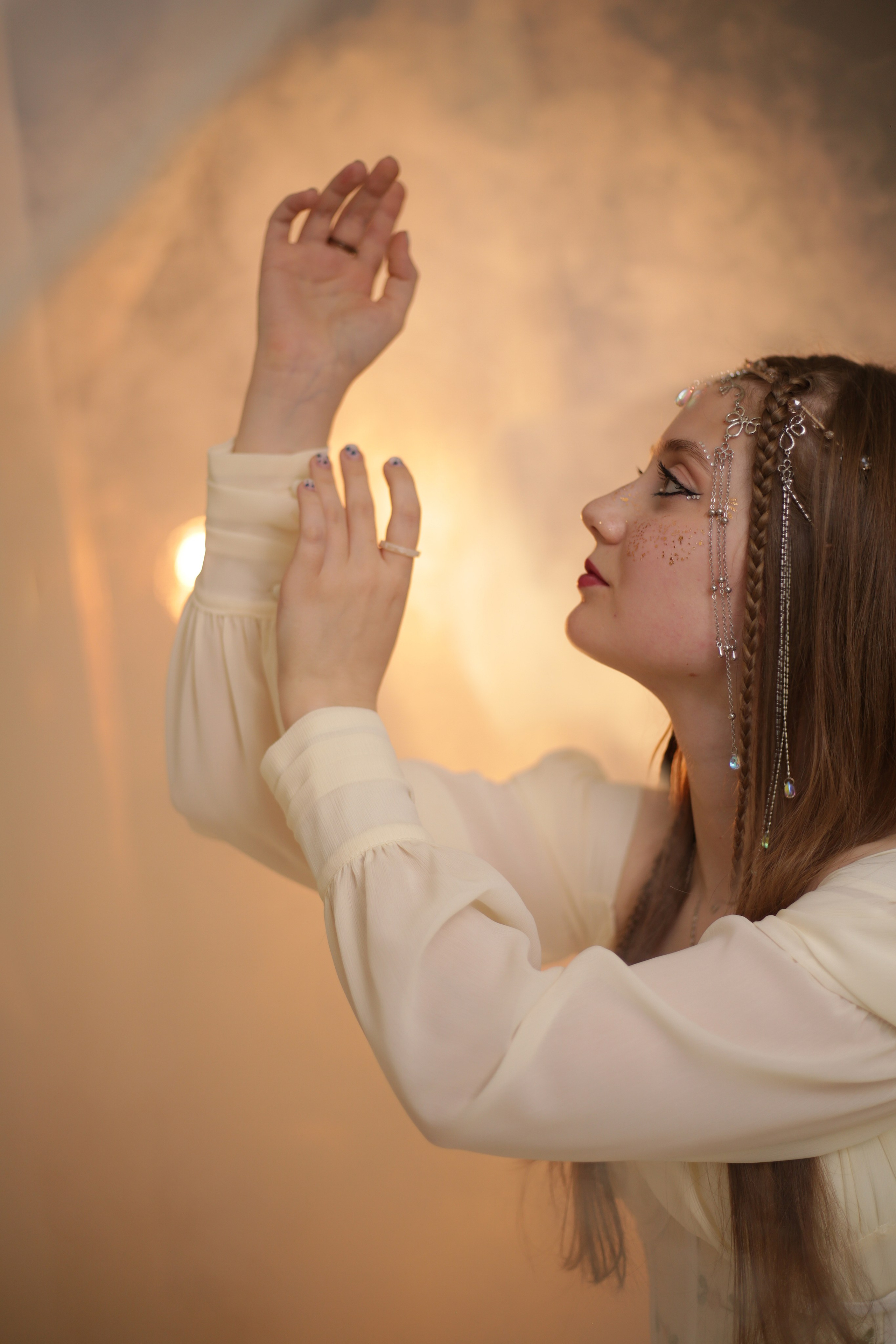 Эльфийская фотосессия: девушка в белом платье с поднятыми руками, лицо украшено блестками, атмосферное освещение