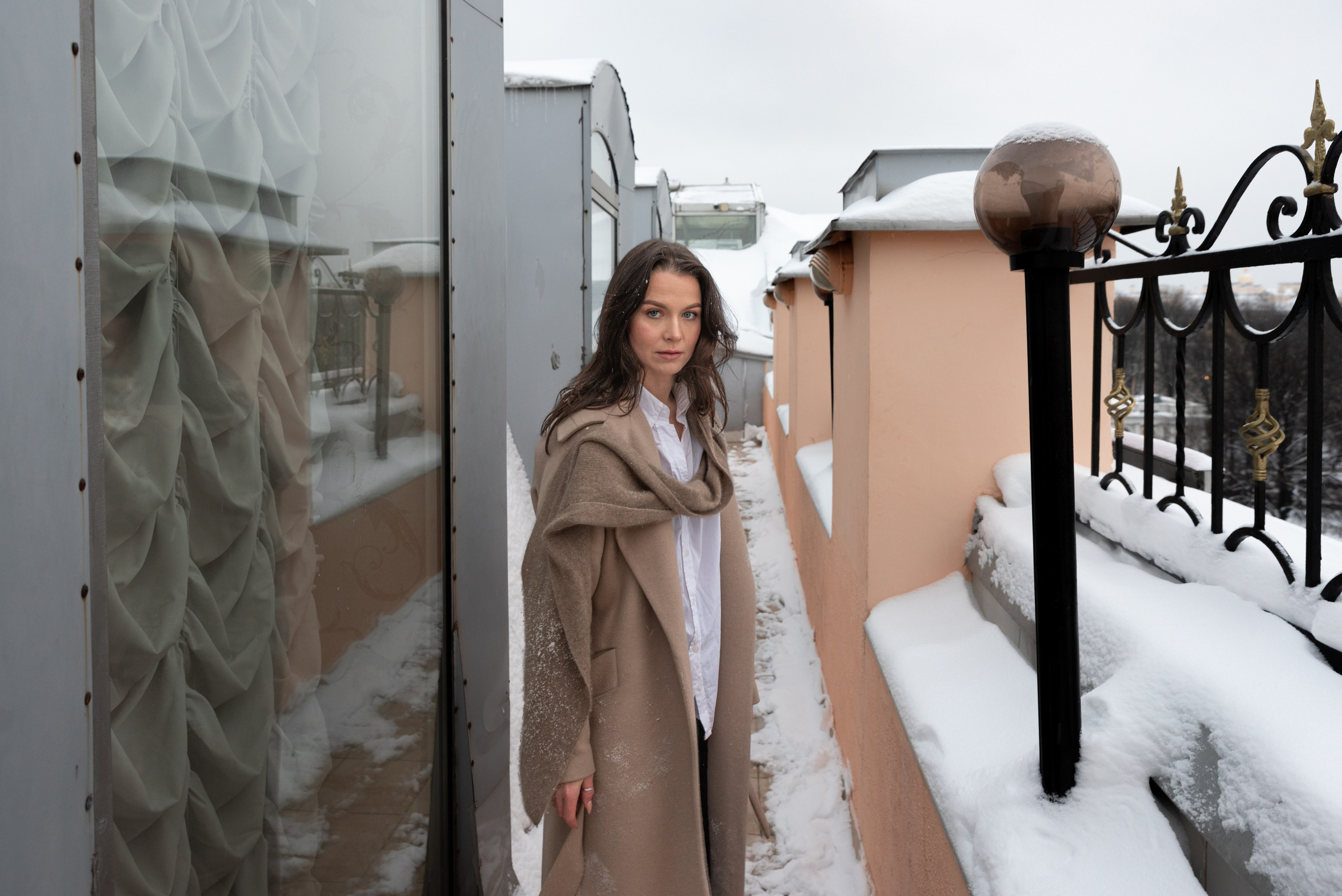 Крыша здания, везде лежит снег. Девушка в бежевом пальто стоит в узком проходе слева высокие окна, справа ограждение и круглый фонарь.