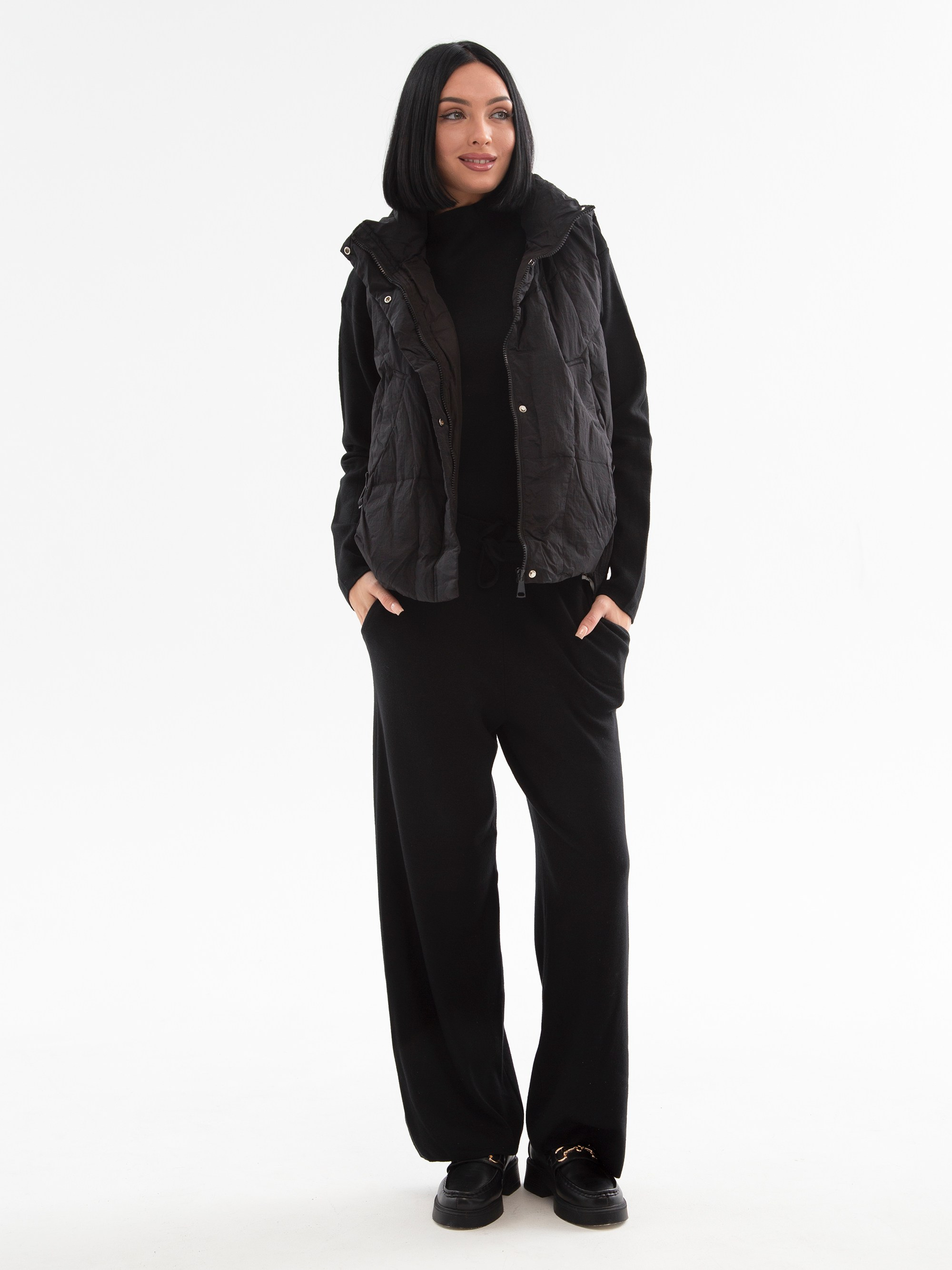 Девушка брюнетка в черном жилете и трикотажном костюме стоит на белом фоне