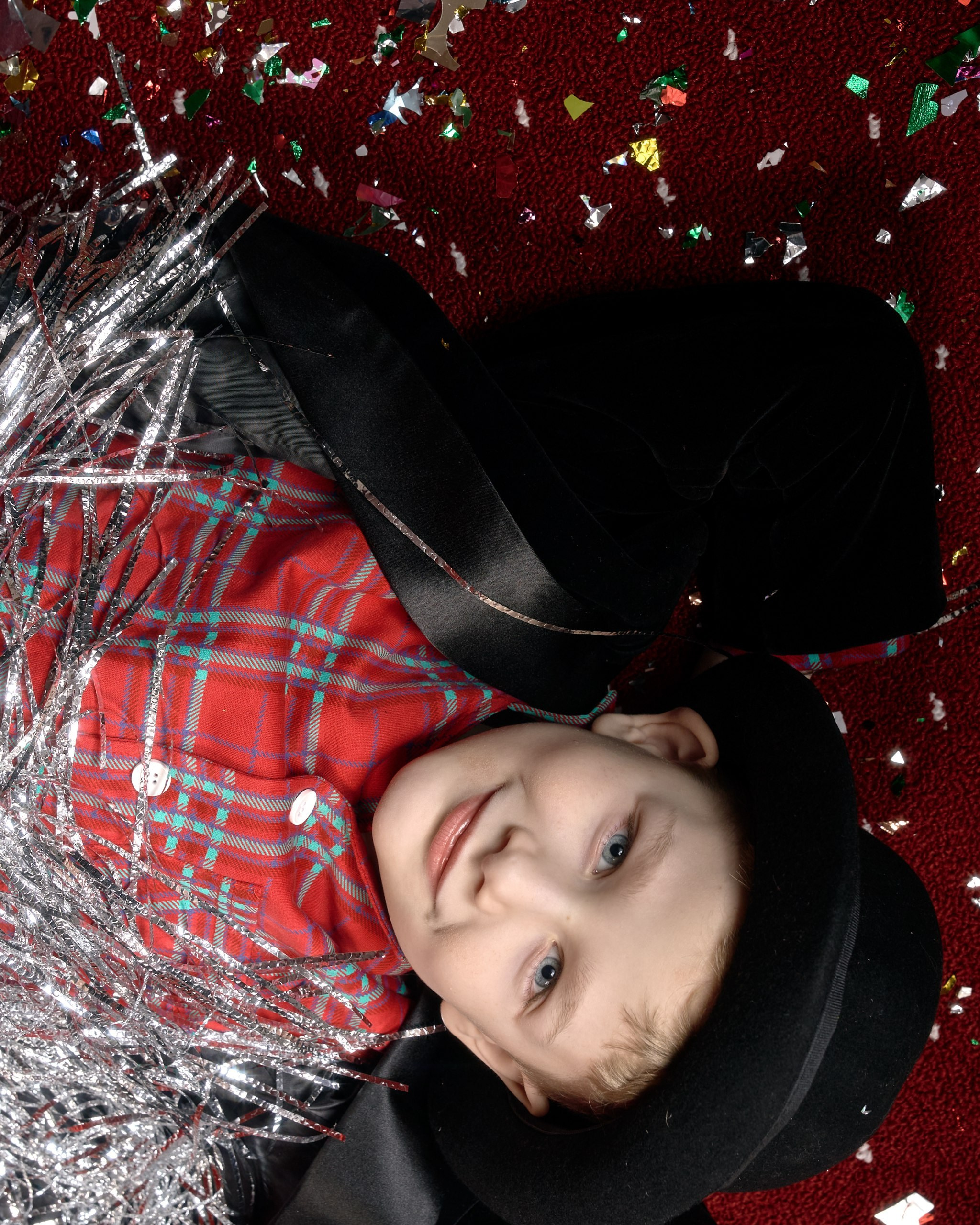 Портрет улыбающегося мальчика в костюме актера: черная шляпа, пиджак и серебряная гирлянда на фоне красного ковра с конфетти. Профессиональная студийная съемка от семейного фотографа. 