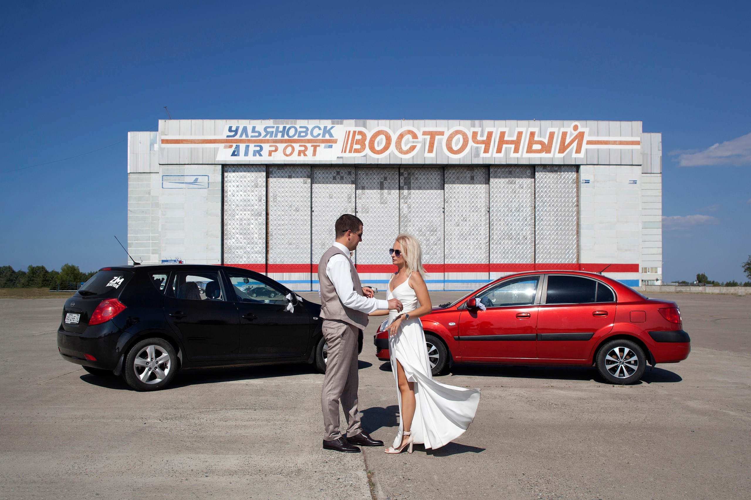 Невеста с женихом стоят на фоне красной и черной машин и здания аэропорта, свадебная фотопрогулка
