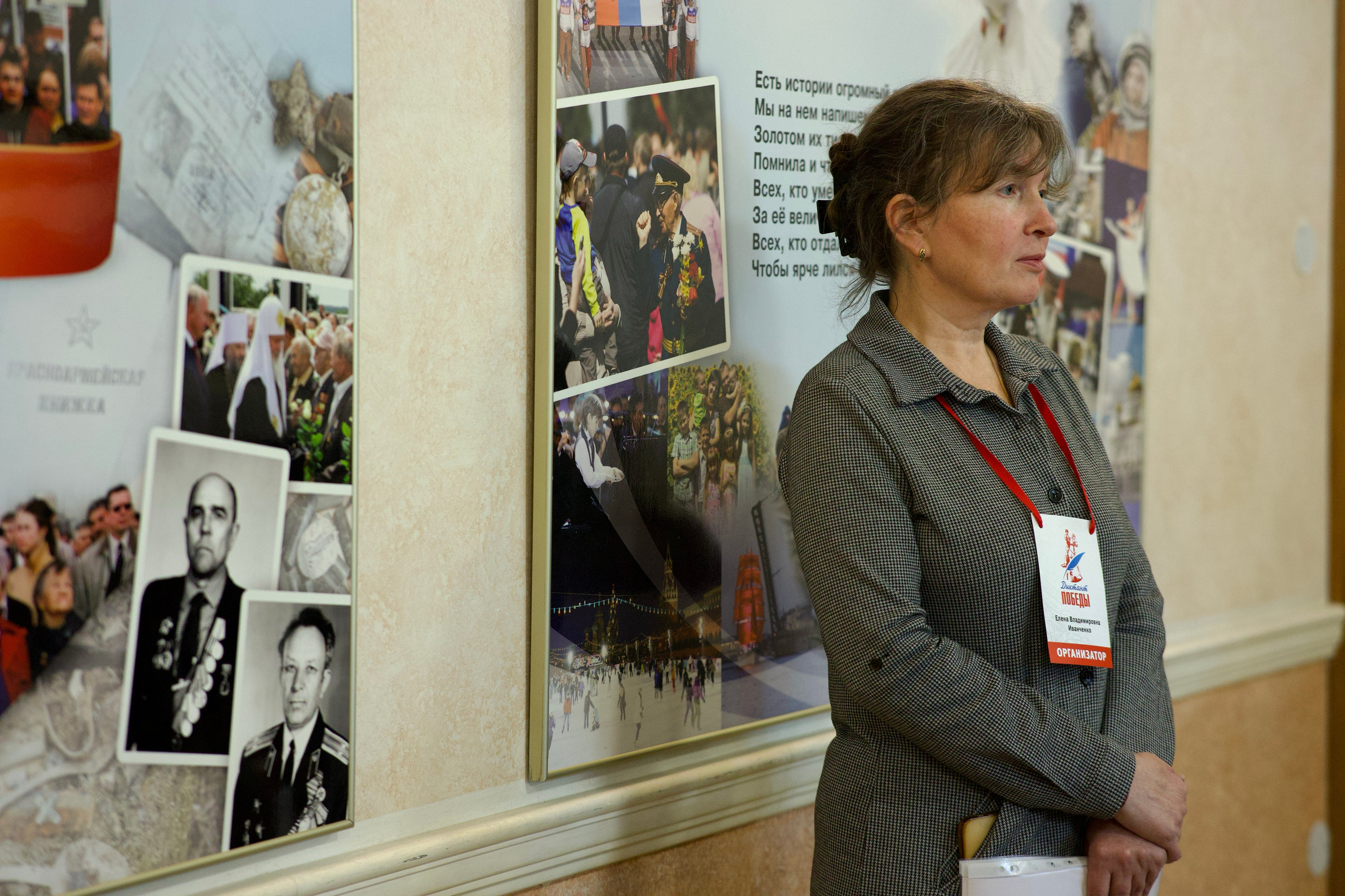 Женщина-организатор в деловом костюме с бейджем стоит в профиль у стенда с архивными фотографиями и текстом стихотворения.