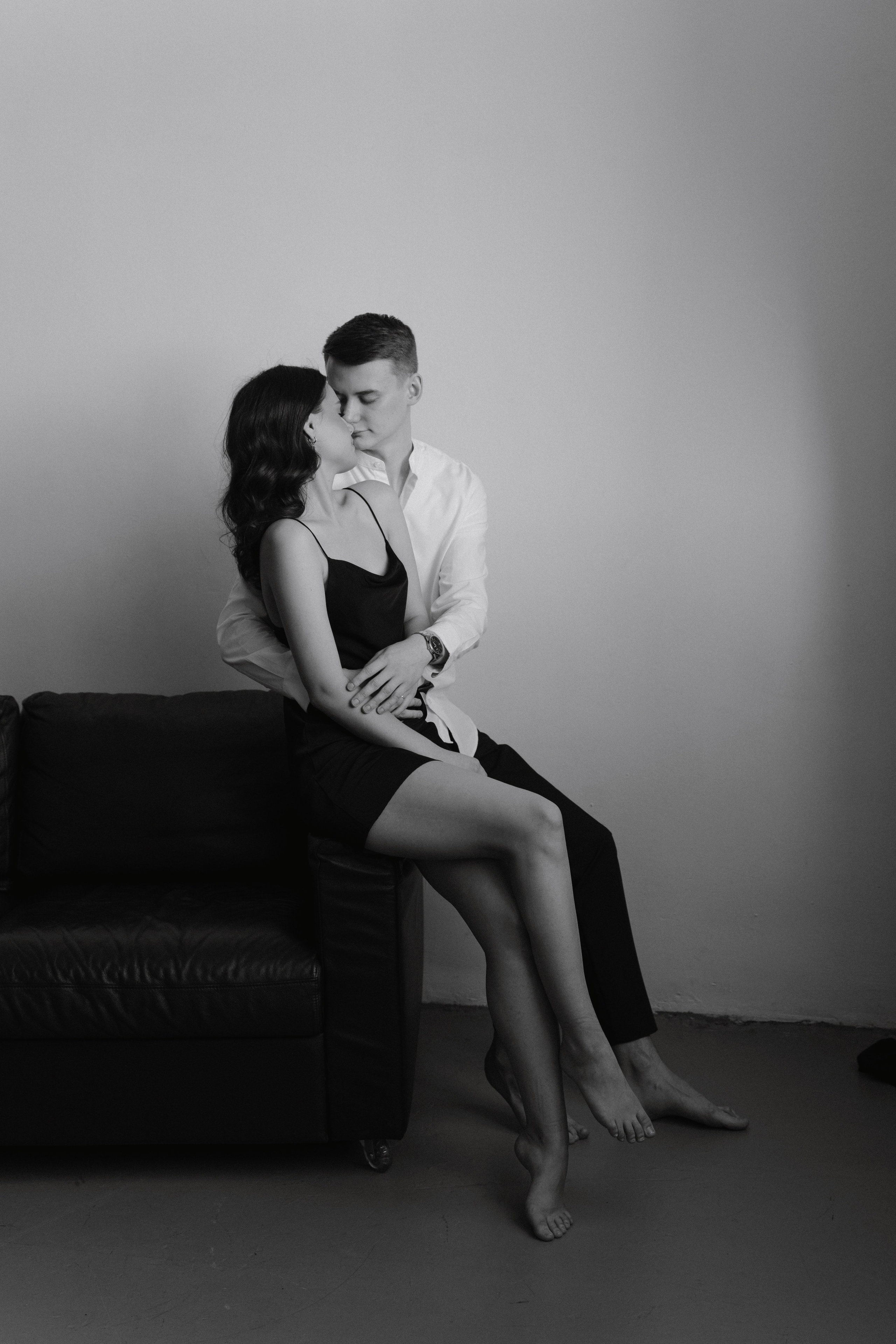 Девушка с парнем на фотосессии лавстори в коротком черном платье сидит на диване