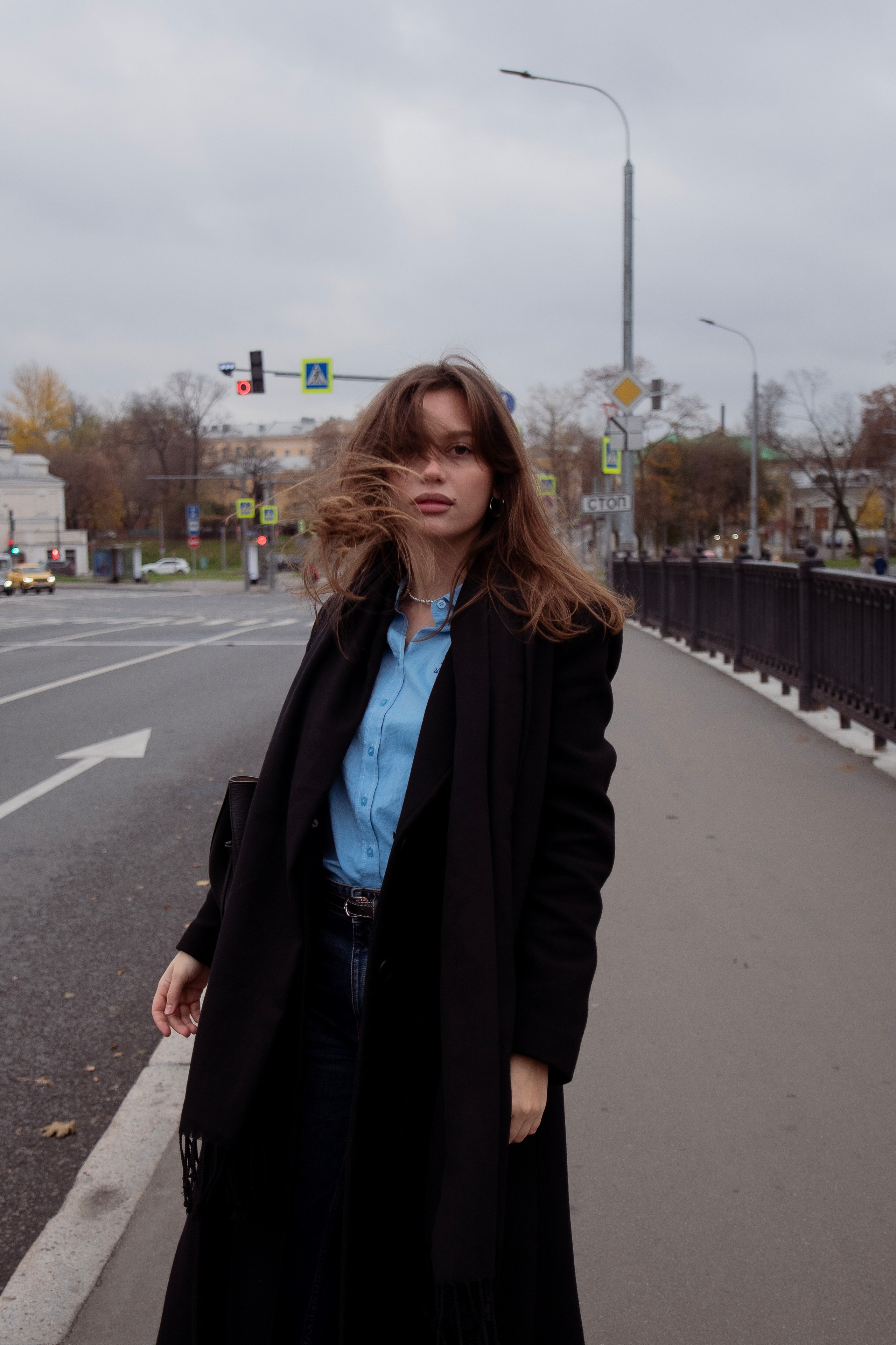 Модель в длинном тёмном пальто и голубой рубашке идёт по городу на фотоне дороги. Уличная fhashionсъемка. Фотосессия на улице. Лучшие локации для фотосессий в Москве.