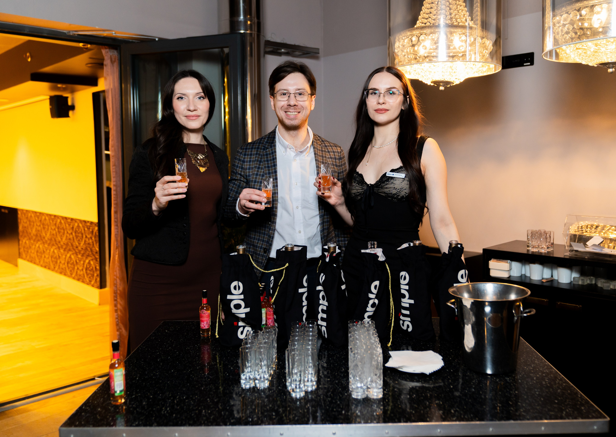 Мужчина и две девушки с коктейлями позируют у барной стойки с подарочными пакетами Simple на вечере в Radisson Sonya. Фото Евгения Мальцева.