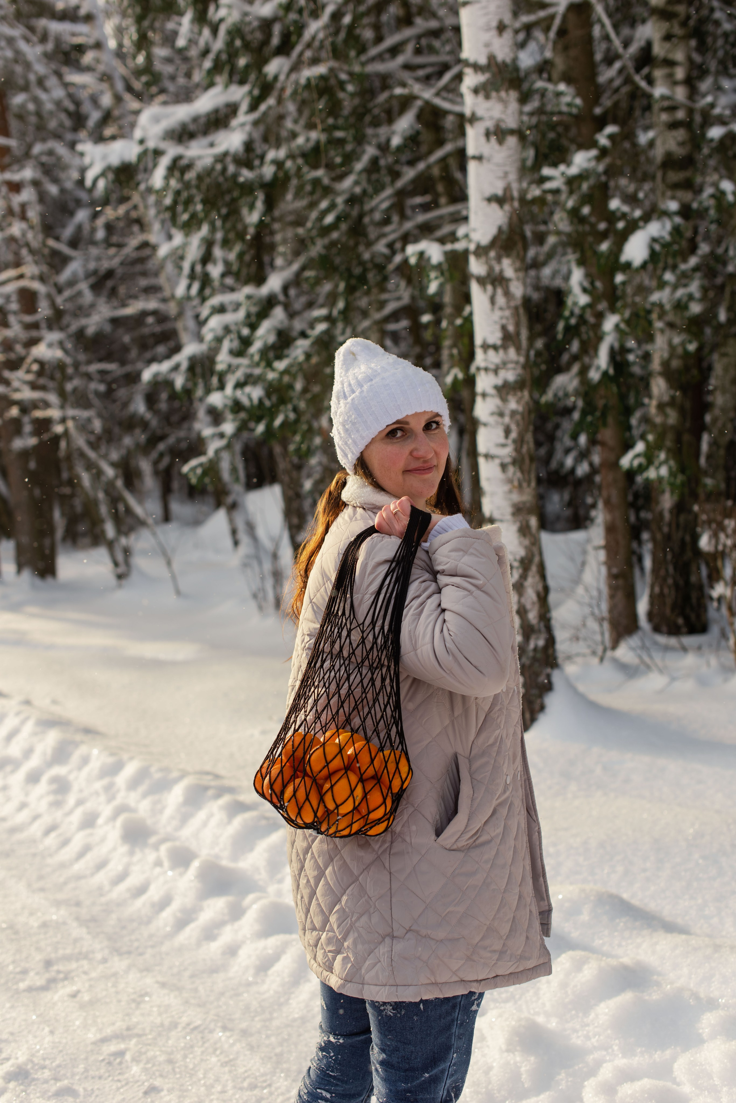 Праздничная фотопрогулка: девушка держит сетку мандарин в зимнем лесу.