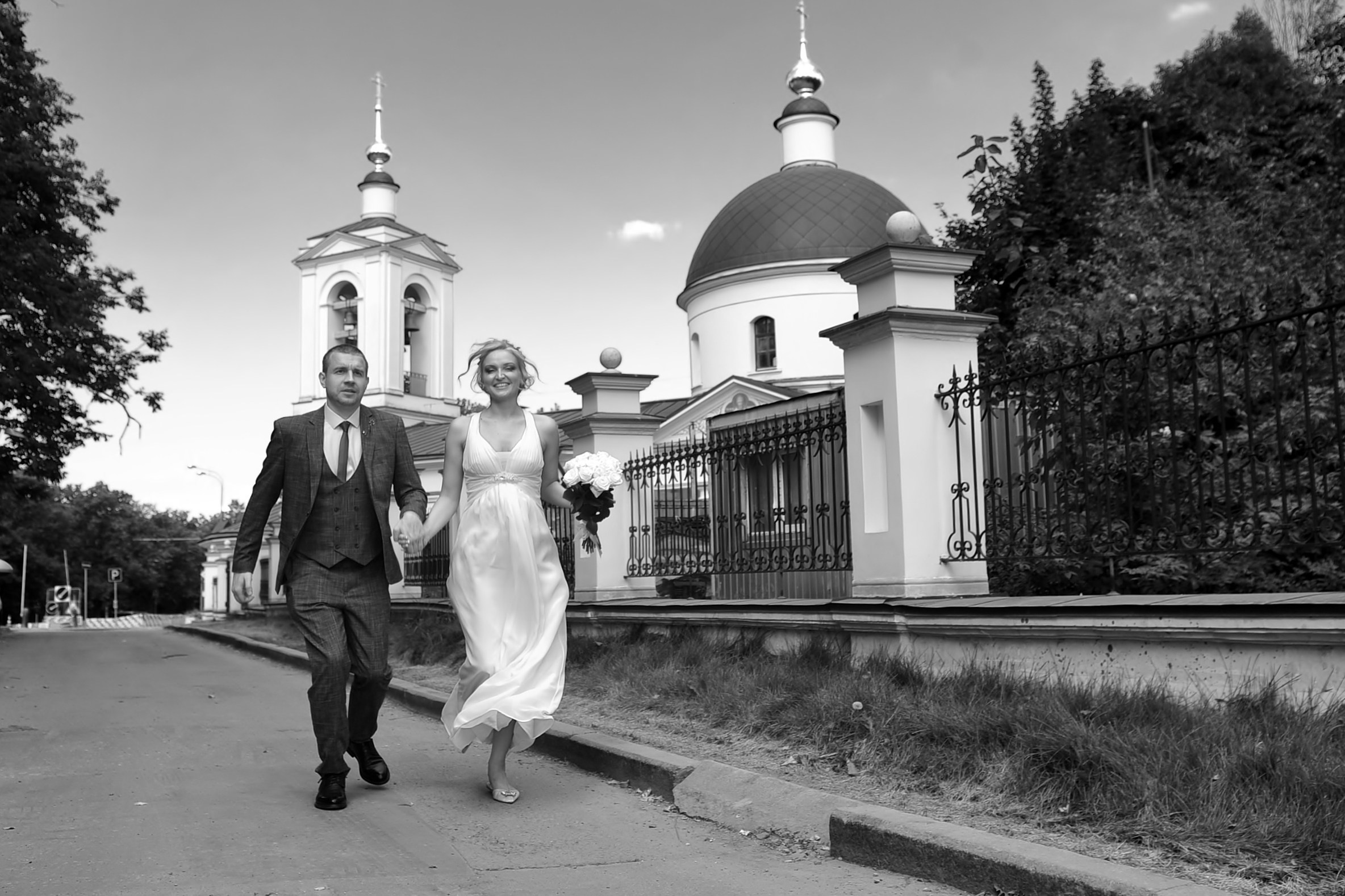Свадьба на Воробьёвых Горах и МГУ, новобрачные идут по дороге вдоль храма и держатся за руки, черно-белая фотография