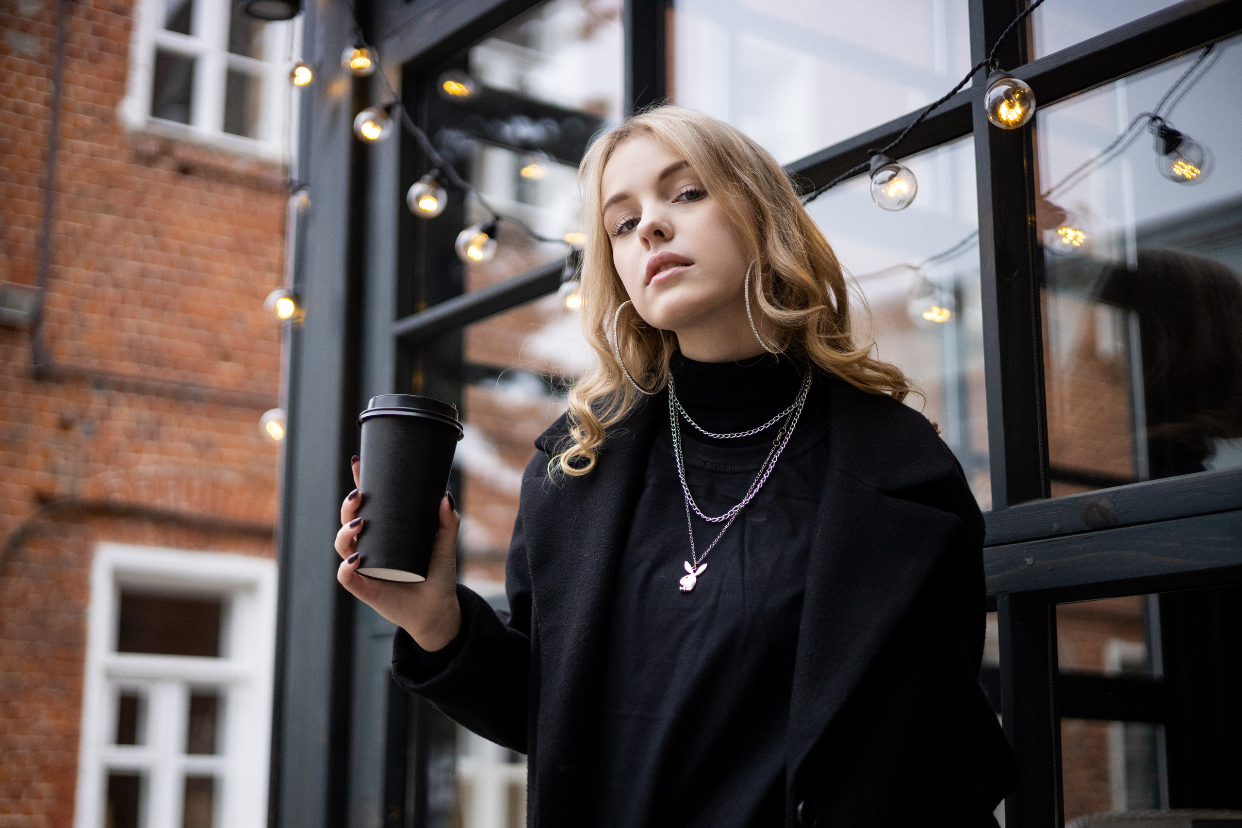 Ученица в чёрном пальто со стаканчиком кофе в руке рядом с кафе.