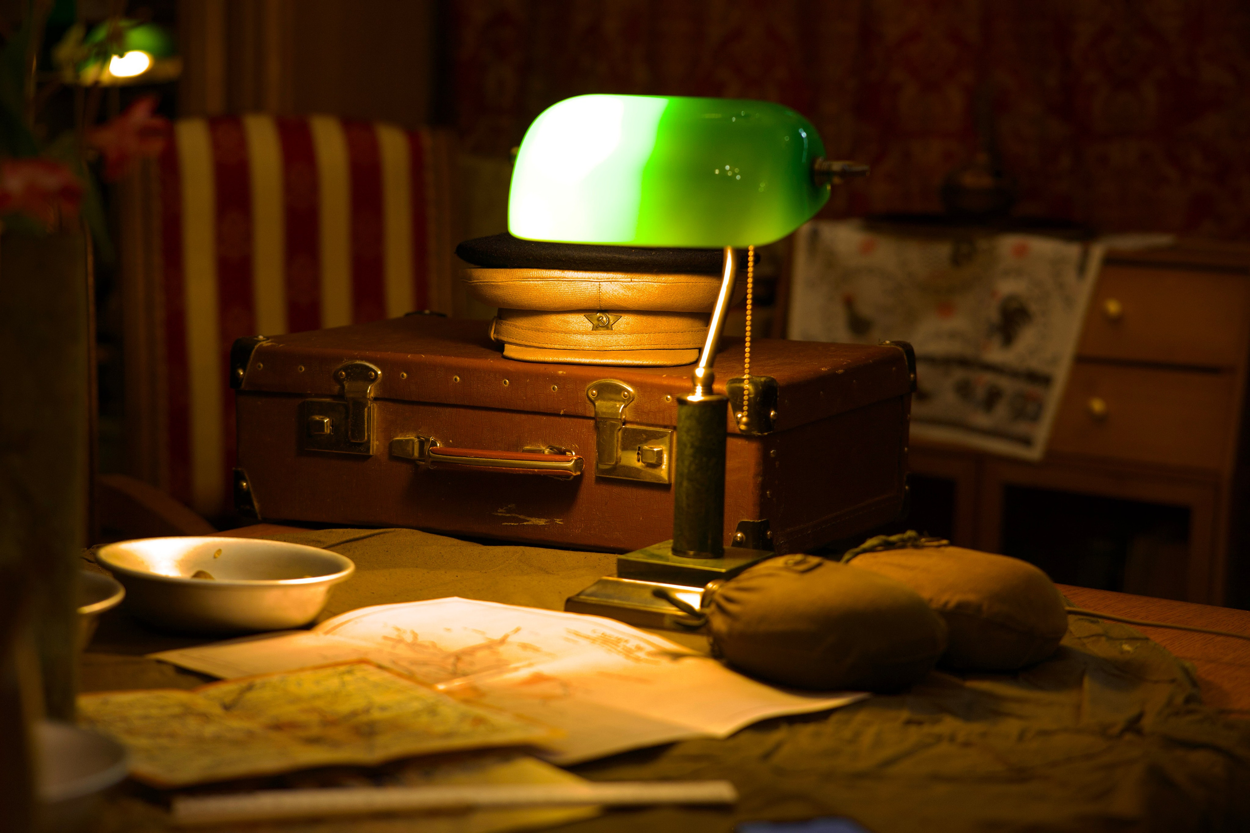 Стол с зелёной банкерской лампой, коричневым кожаным чемоданом и военной фуражкой. На столешнице — карта и пепельница. Тёплый вечерний свет.