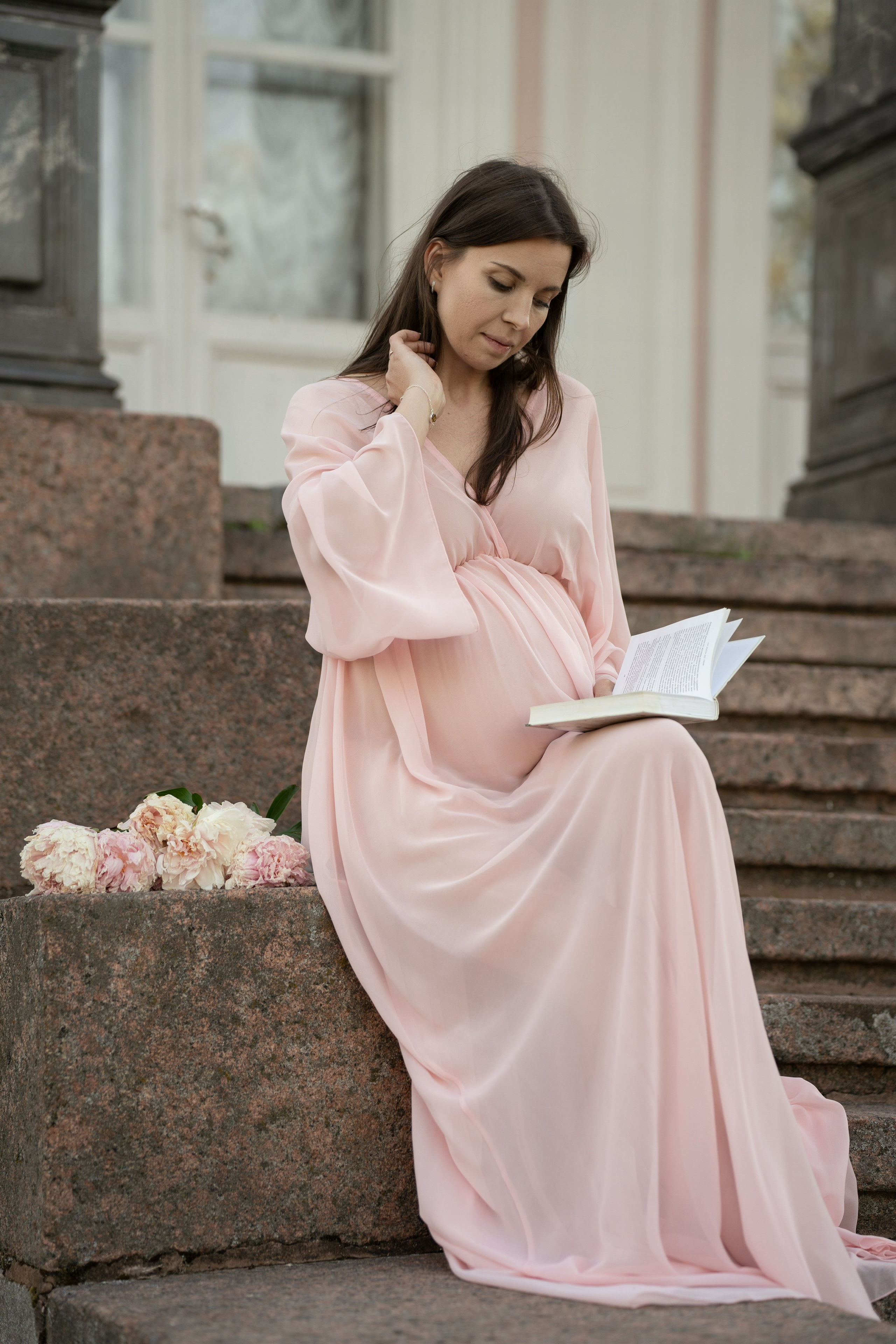 Будущая мама читает книгу на ступеньках заброшенного особняка.