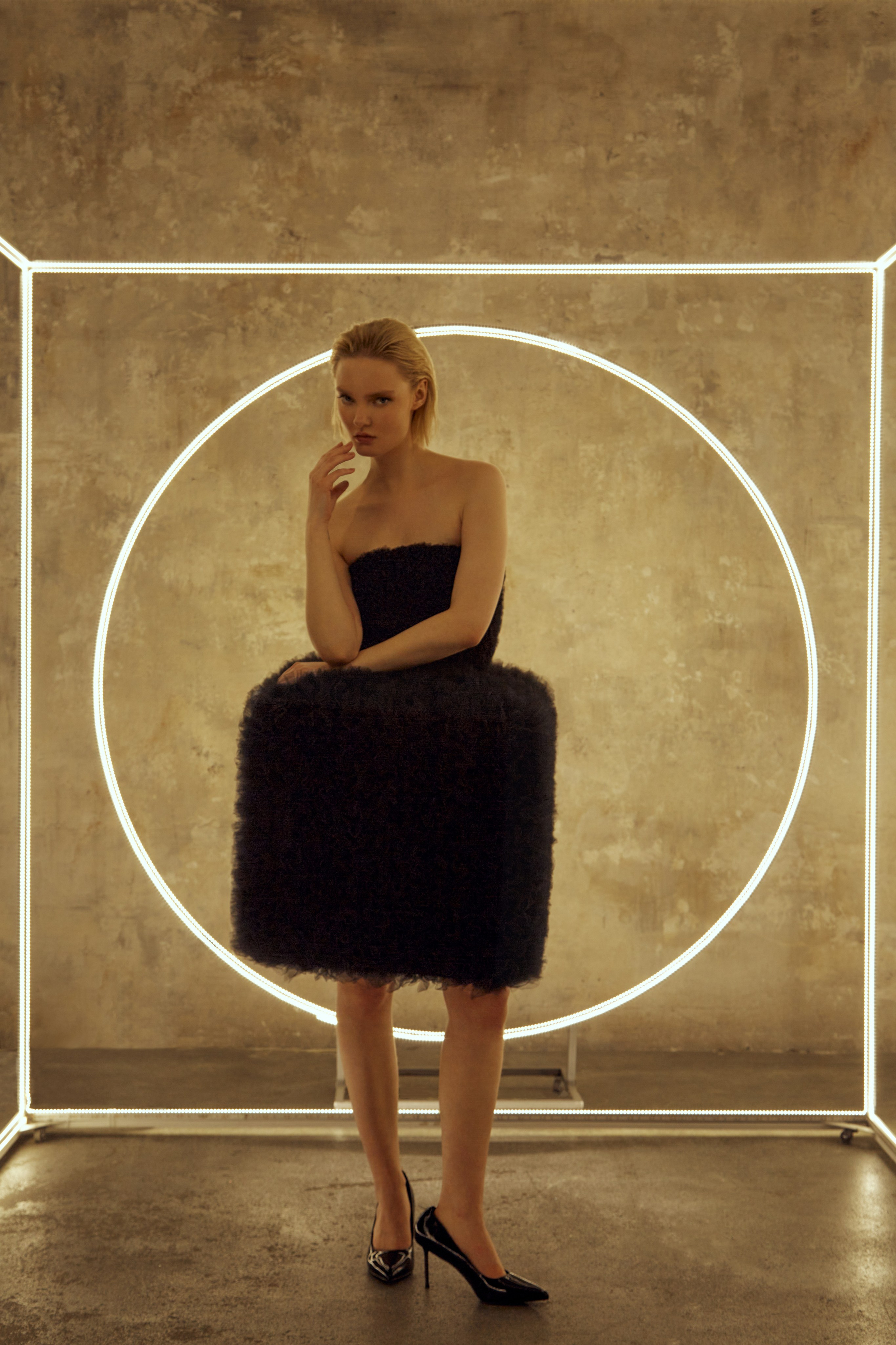 Девушка в черном объемном платье и черных туфлях на высоком каблуке стоит на фоне светящегося куба