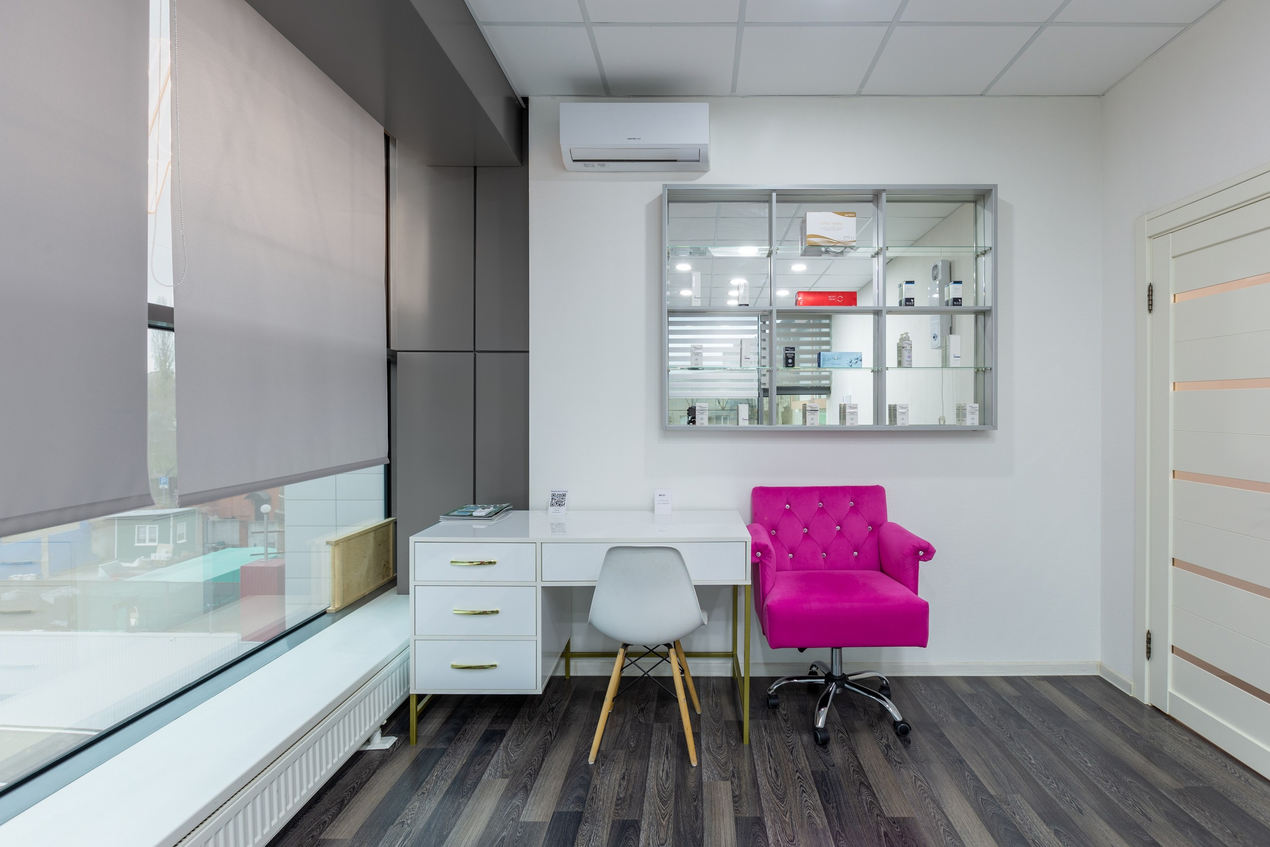 Интерьерное фото кабинета косметолога. Розовое мягкое кресло для клиента. Рабочий стол у окна в интерьере.