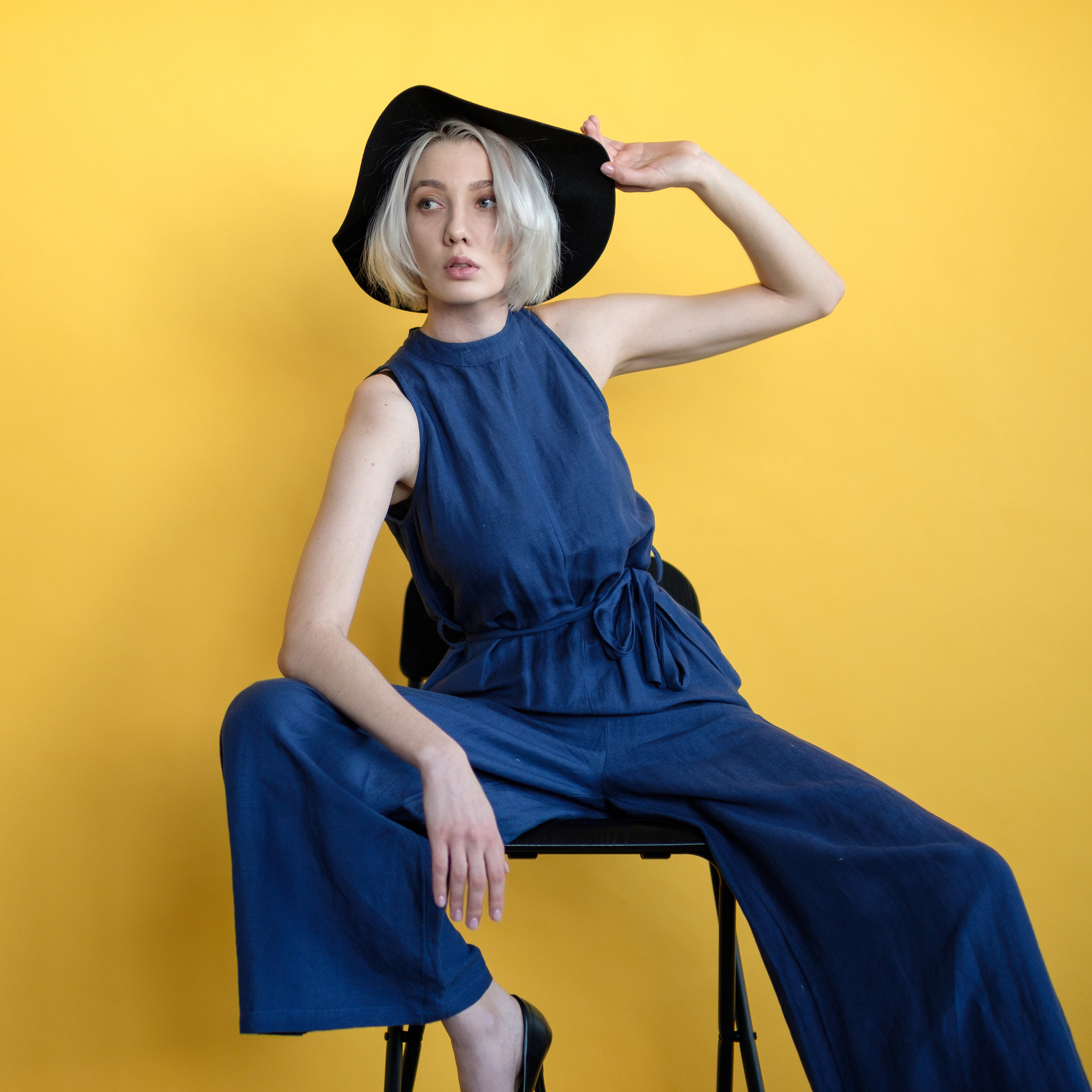 Девушка в синем брючном костюме позирует,сидя на стуле на желтом фоне.Держит одной рукой шляпу и смотрит в сторону от фотографа.