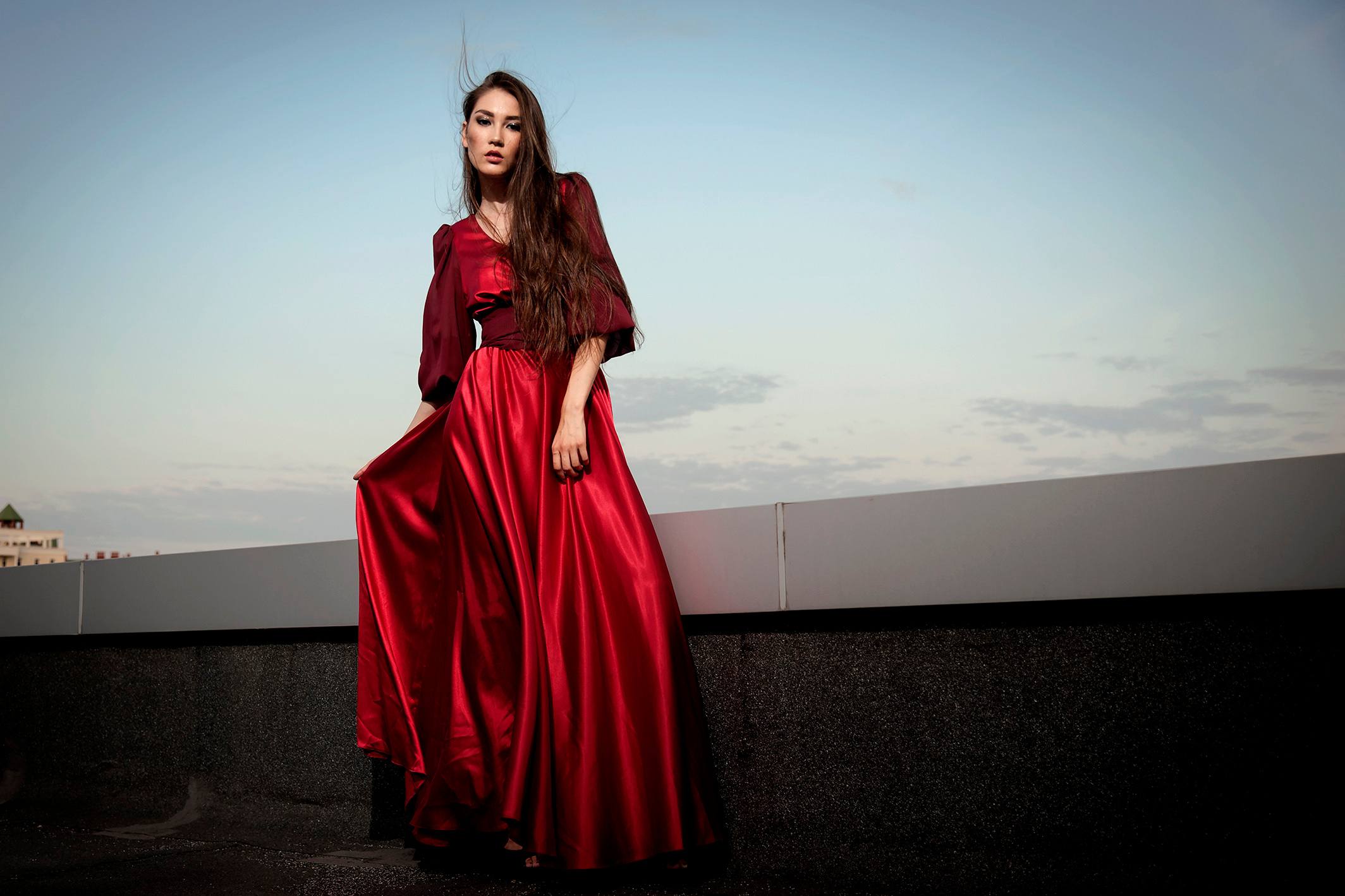 Красивая девушка в ярко-красном платье стоит на крыше здания.Ветер развевает ее волосы.