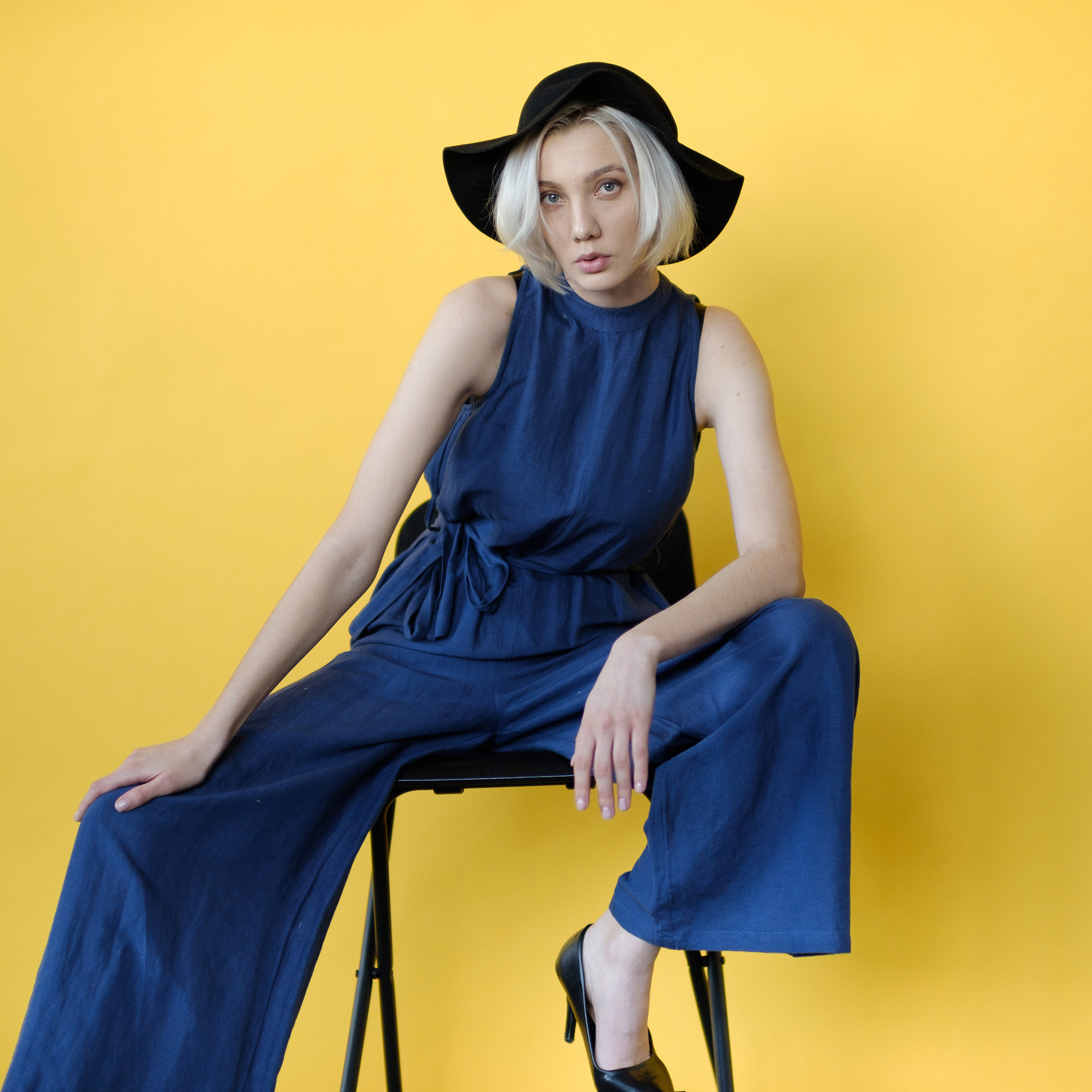 Красивая блондинка в синем брючном костюме позирует на желтом фоне,сидя на стуле.