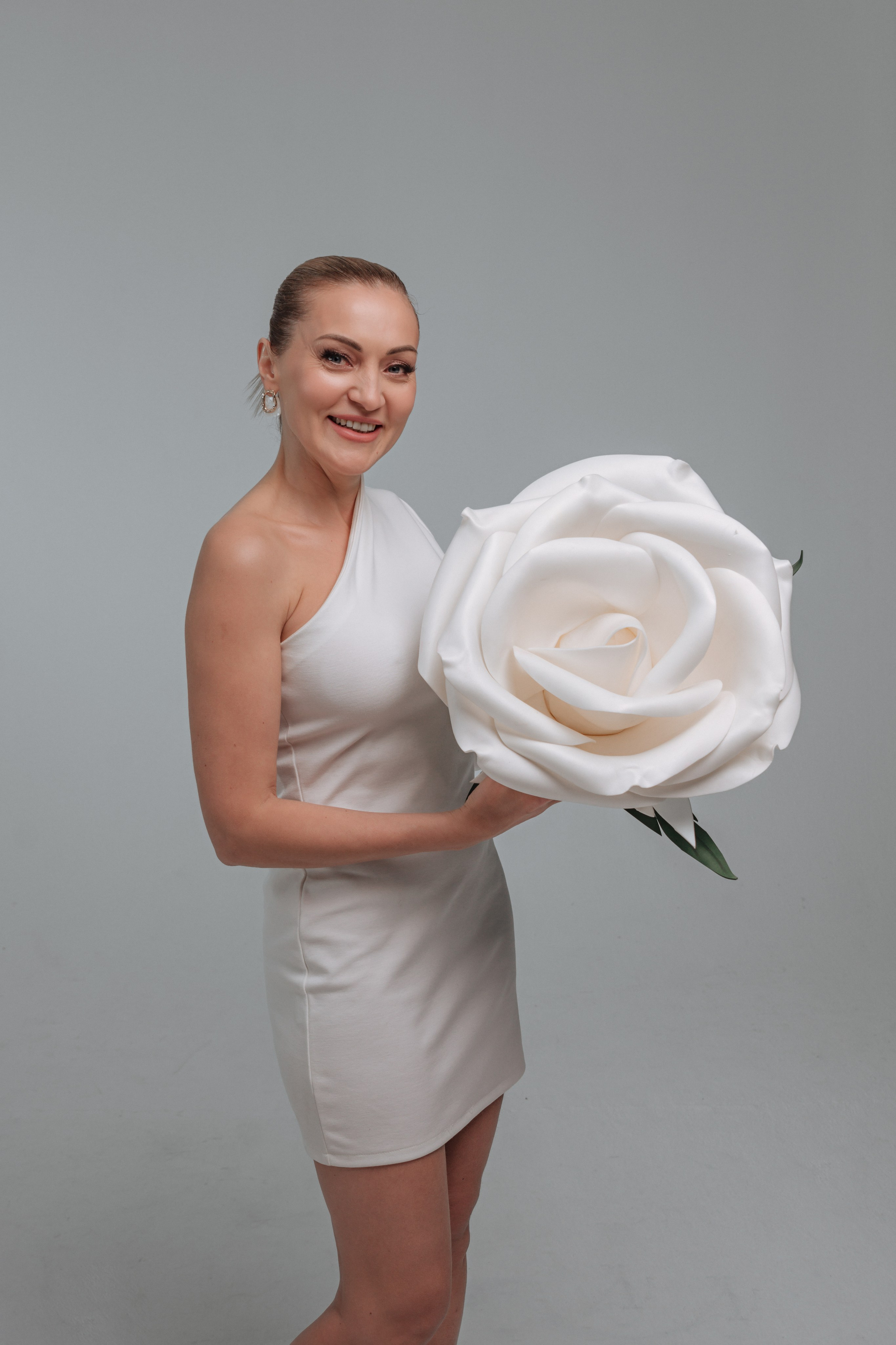 На переднем плане изображена молодая женщина, одетая в элегантное белое платье. Платье струится мягкими складками, подчеркивая ее стройную фигуру. Ткань переливается на солнце, создавая ощущение легкости и воздушности.