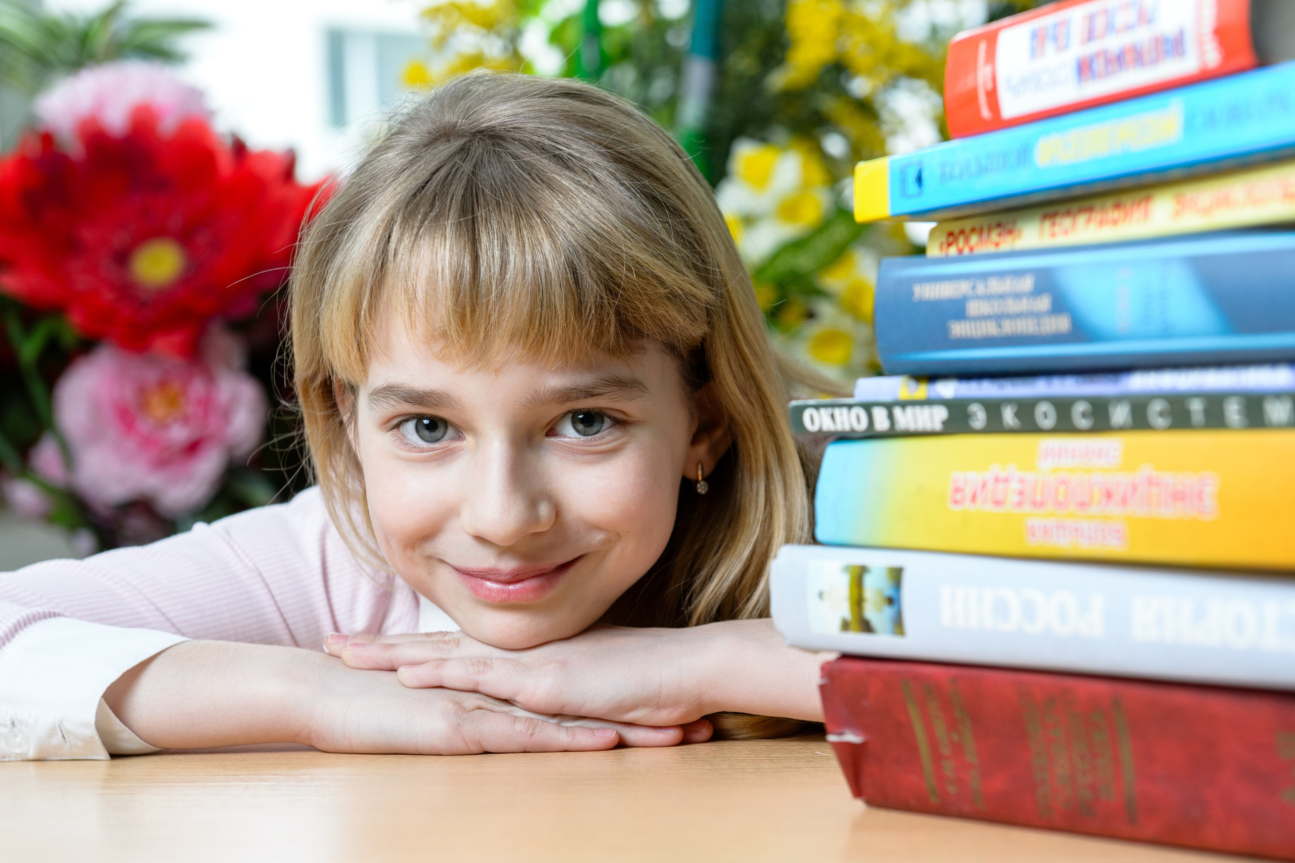 Школьница улыбается положив голову на руки, рядом сложена высокая стопка разноцветных книг.