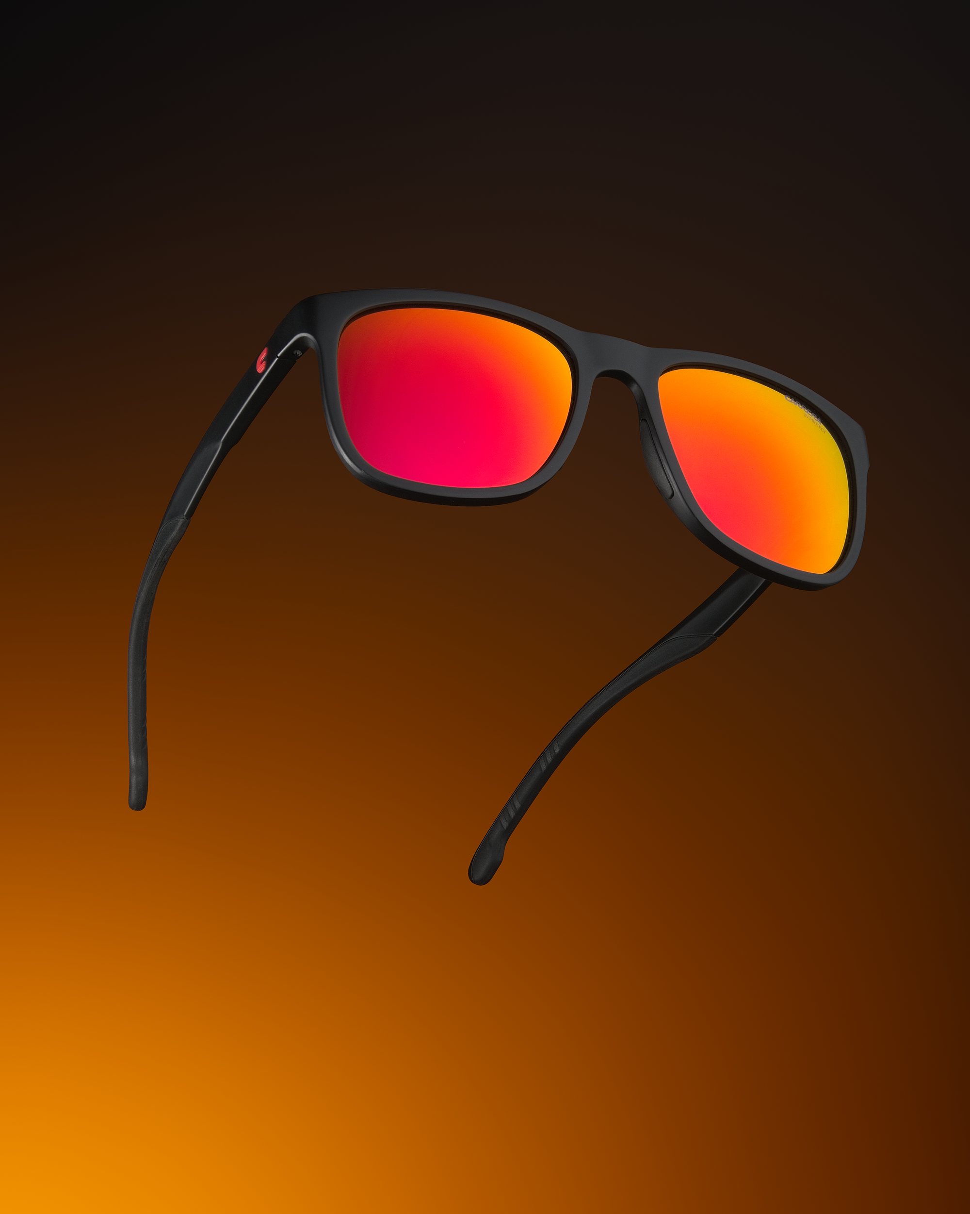 Студийная фотография черных матовых солнцезащитных очков с ярко-красными и оранжевыми зеркальными линзами. Очки изображены в парящем положении на эффектном градиентном фоне, переходящем от теплого оранжевого к глубокому черному, подчеркивая динамику и современный стиль.