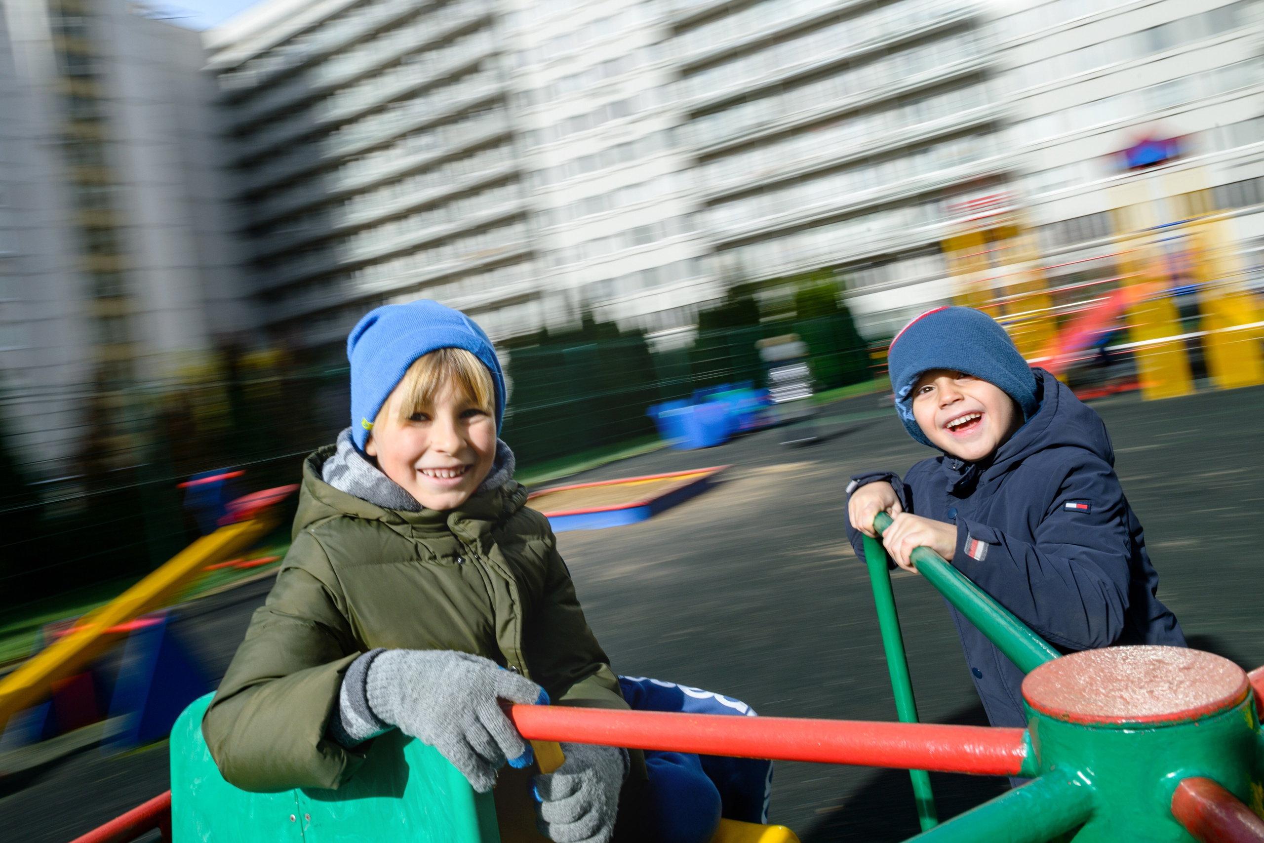Два мальчика в шапках катаются на карусели на уличной площадке, кадр с динамичным размытием фона.