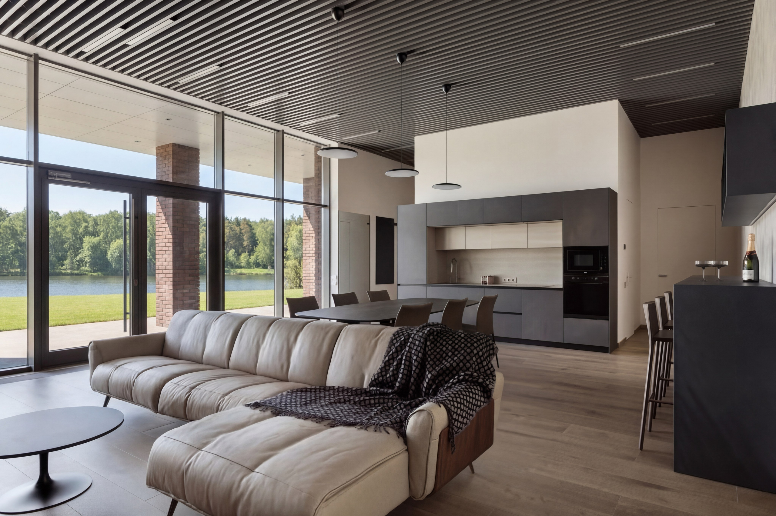 Современная lounge-зона с темной кухней и светлым кожаным диваном от Валерии Котович: реечный потолок, панорамные окна с видом на реку и минималистичный дизайн в премиальном доме у воды.