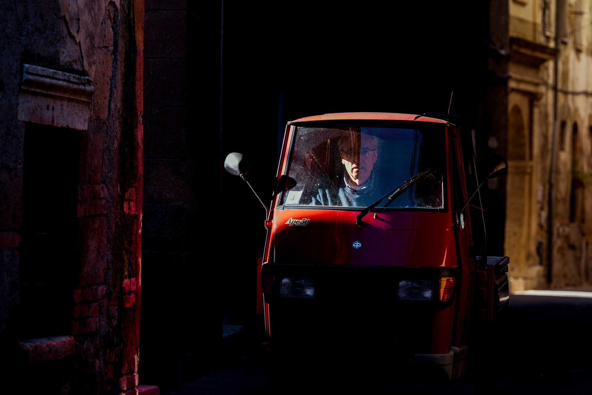 Крупный план красного Piaggio Ape, выезжающего из тени на итальянской улице.