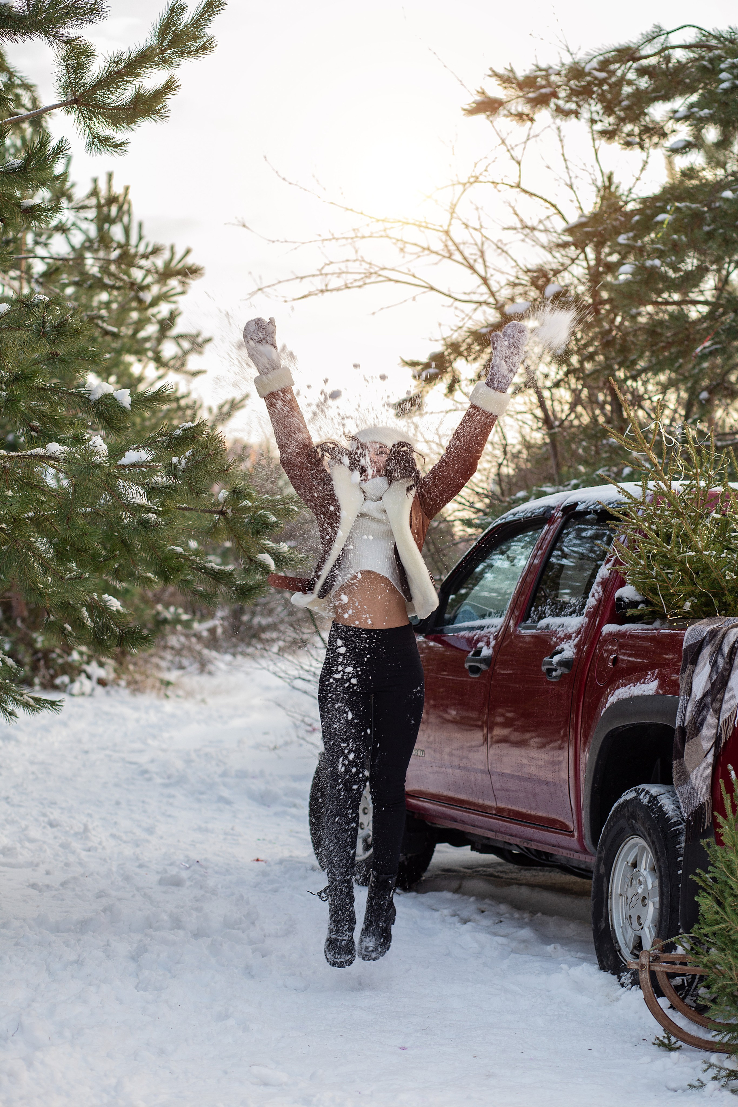 девушка в коричневой  куртке  прыгает, играет со снегом. рядом  стоит красный пикап. зима. снег. закат. 